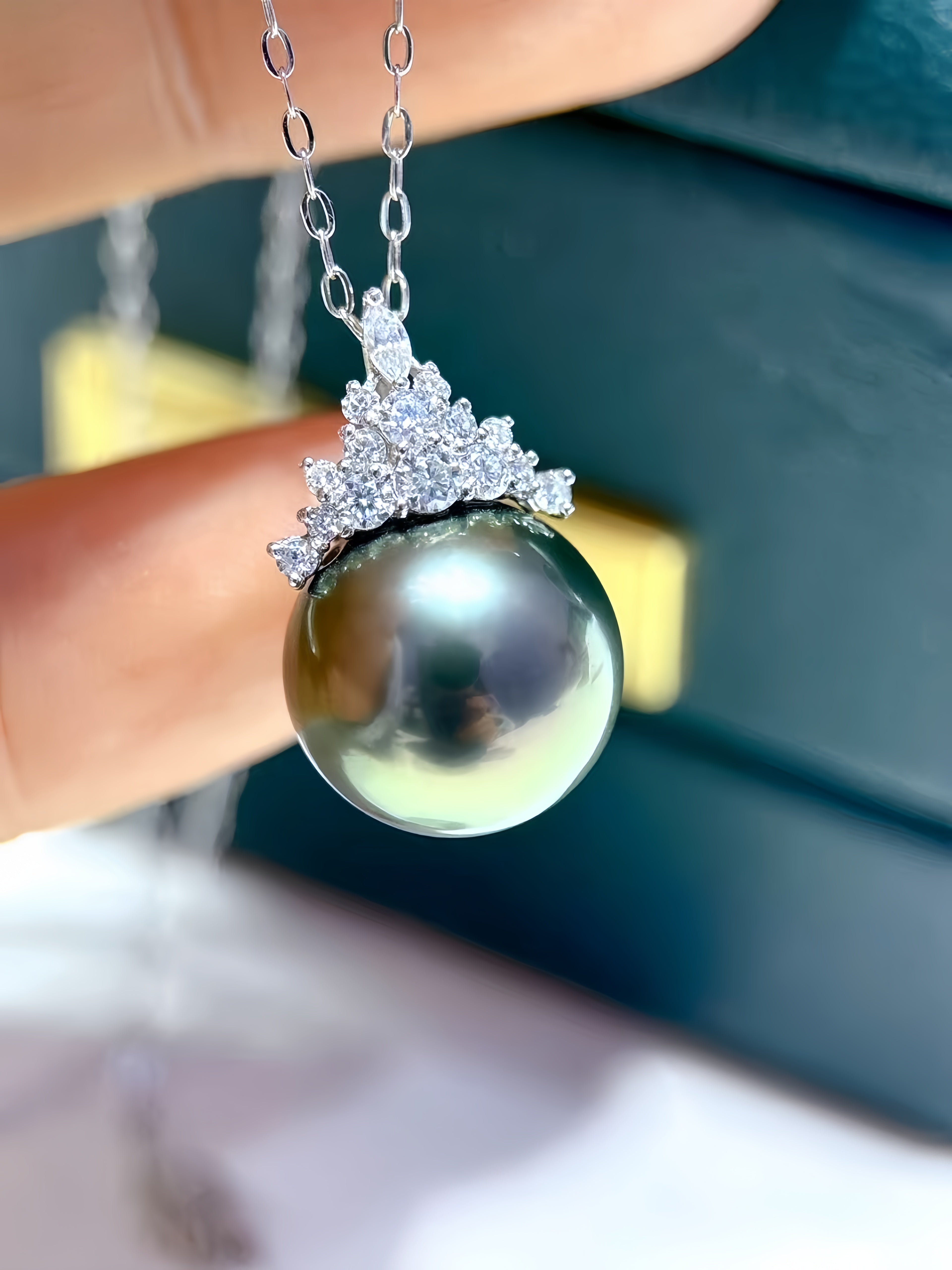 A close-up of a pendant necklace held by fingers, featuring a large round dark pearl pendant set in a silver-toned floral cluster with small clear stones, against a dark blue background.