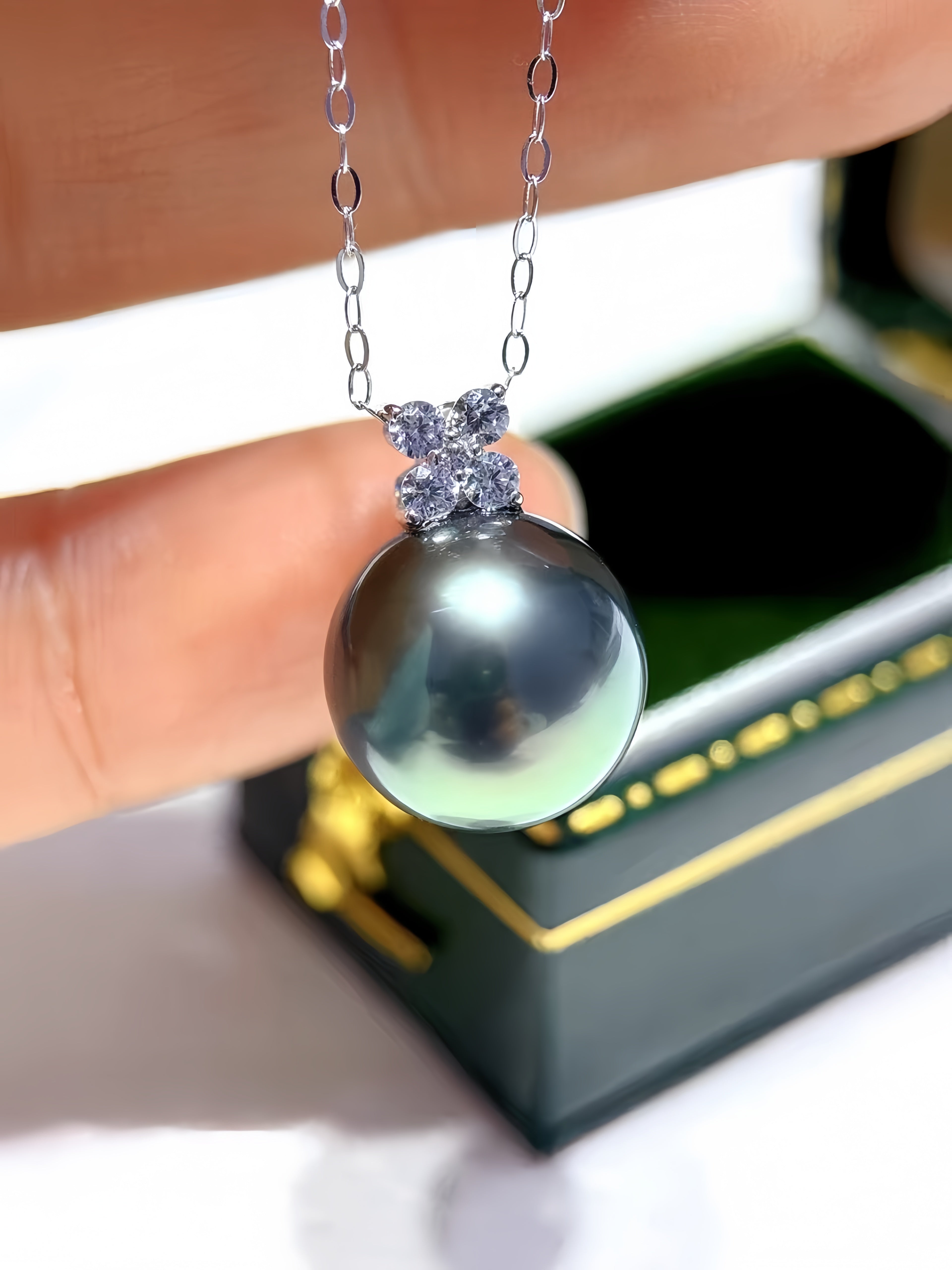 A close-up of a silver pendant necklace featuring a large, round, dark gray pearl with a silver floral halo, held by fingers with a jewelry box in the background.