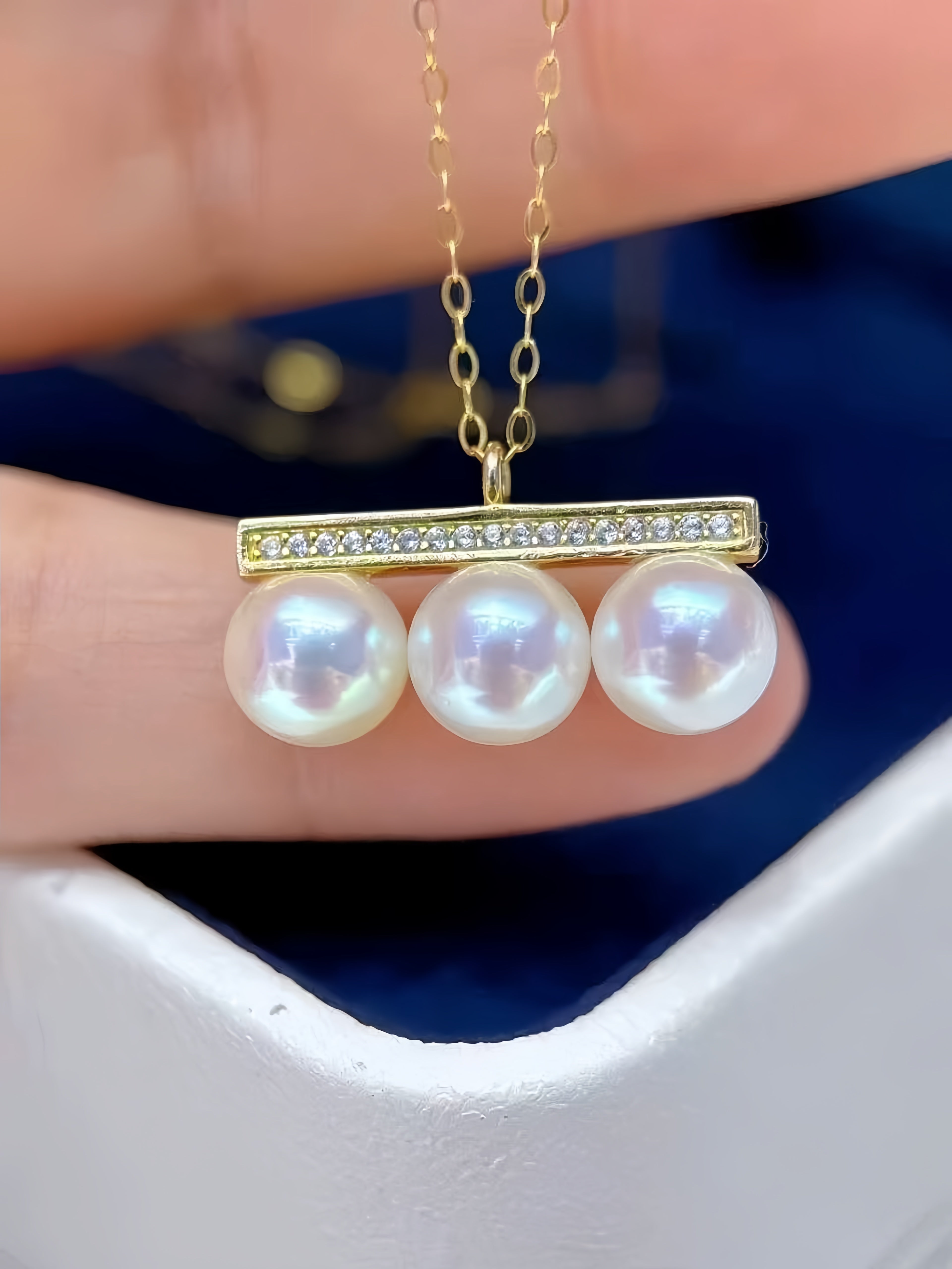 A close-up of a gold necklace with a bar pendant featuring three large white pearls and a row of small clear stones, held between fingers against a dark blue background.