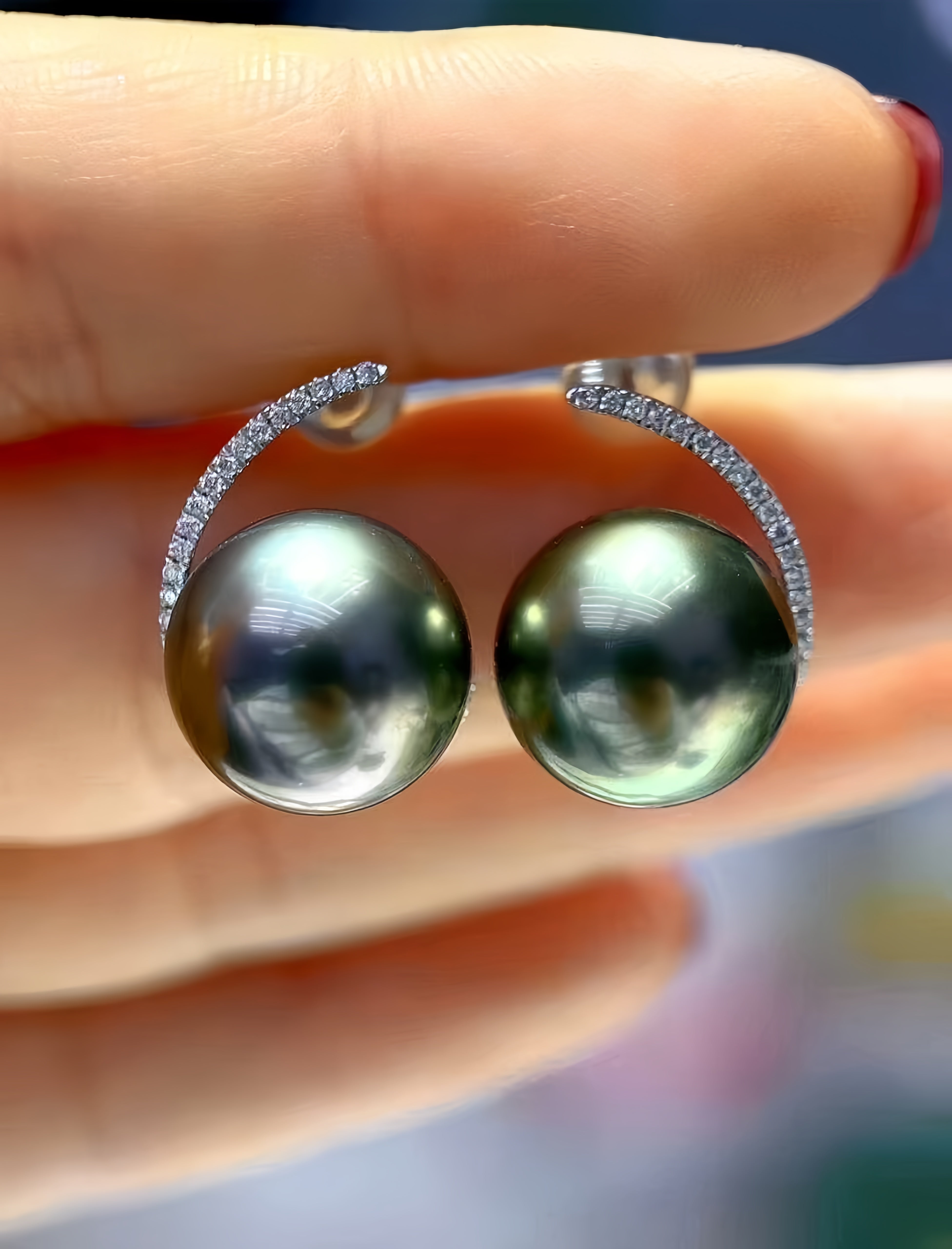 A close-up of a hand holding a pair of earrings with large round dark green pearls and silver settings accented with small clear stones.