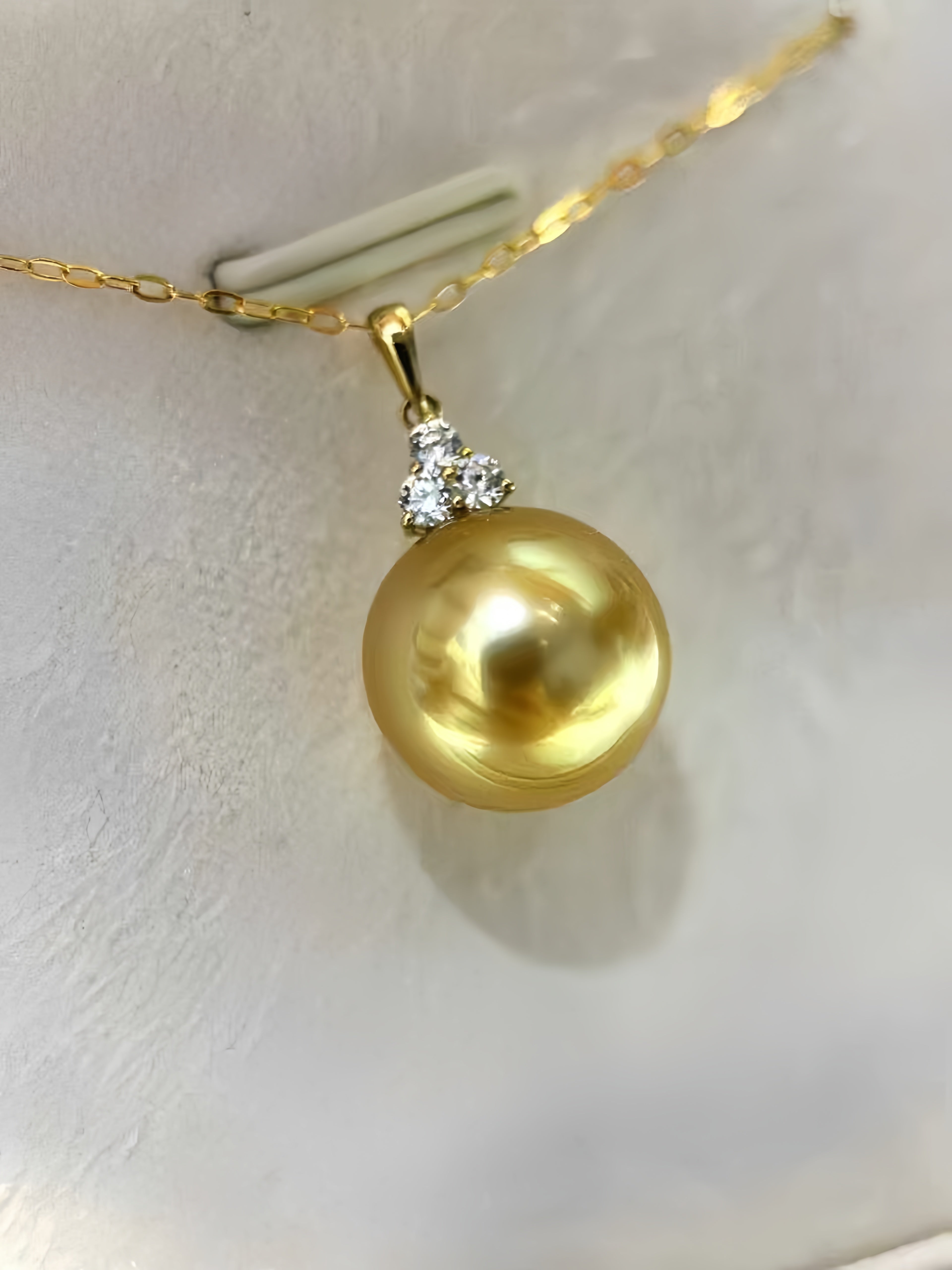 A gold pendant necklace featuring a large, lustrous gold pearl pendant with a small cluster of clear stones at the bail, shown on a gold chain against a textured white background.