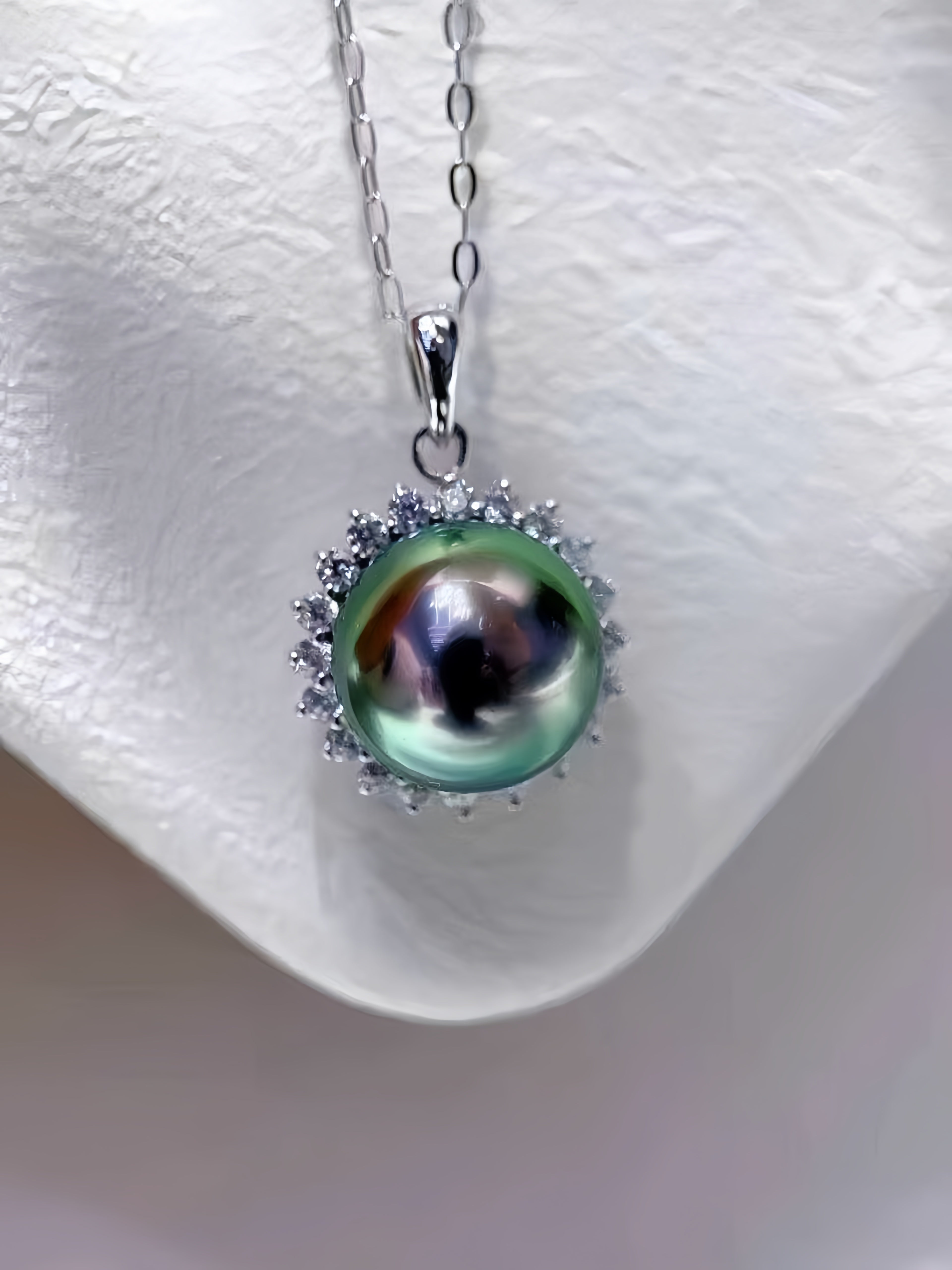 A silver pendant necklace featuring a large, iridescent green pearl surrounded by a halo of small clear stones, displayed on a textured white jewelry bust.