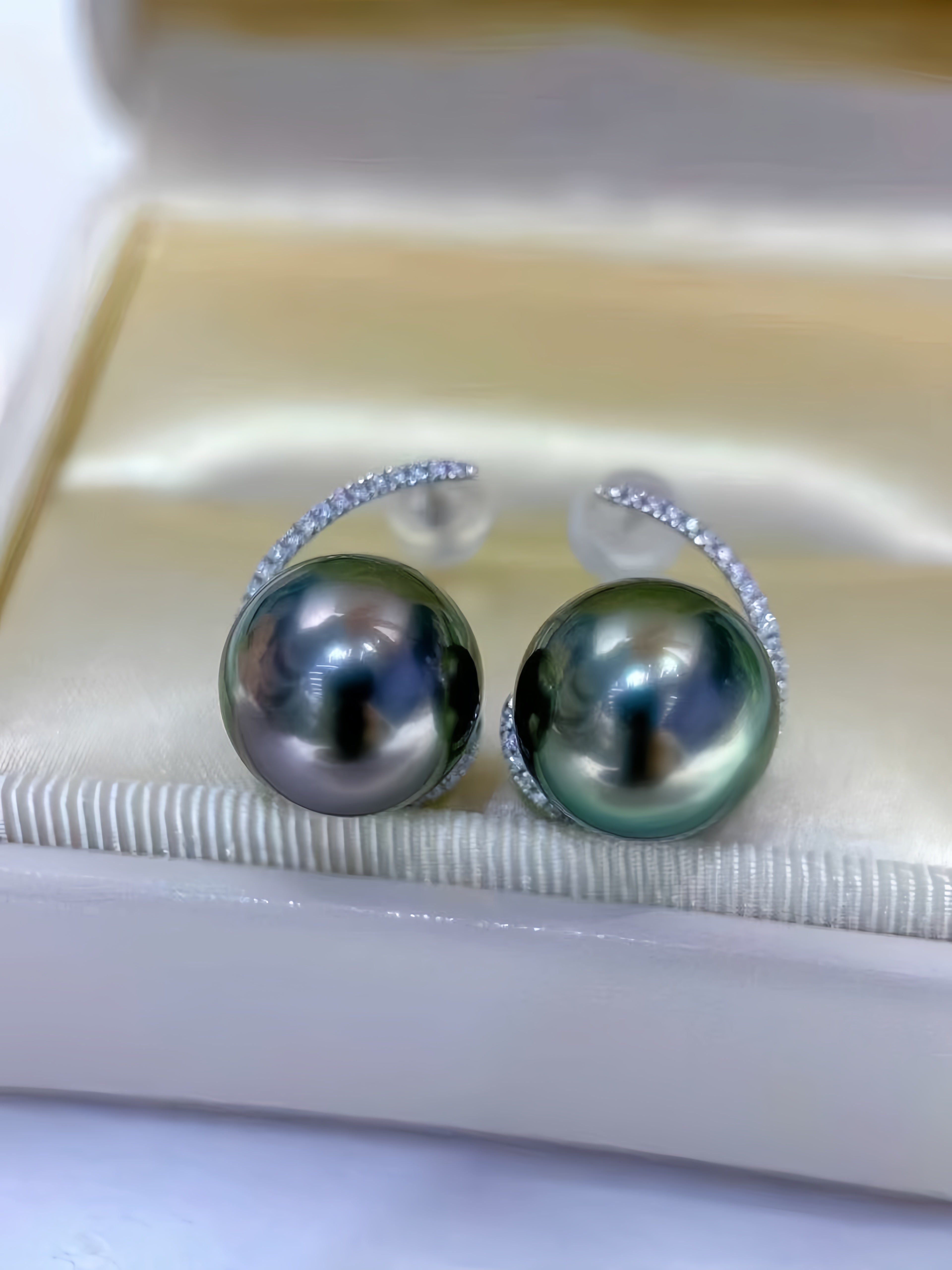 A pair of earrings featuring large round Tahitian pearls with a dark green-gray iridescent sheen, set in a silver-toned crescent shape accented with small clear stones, resting inside a cream-colored jewelry box.