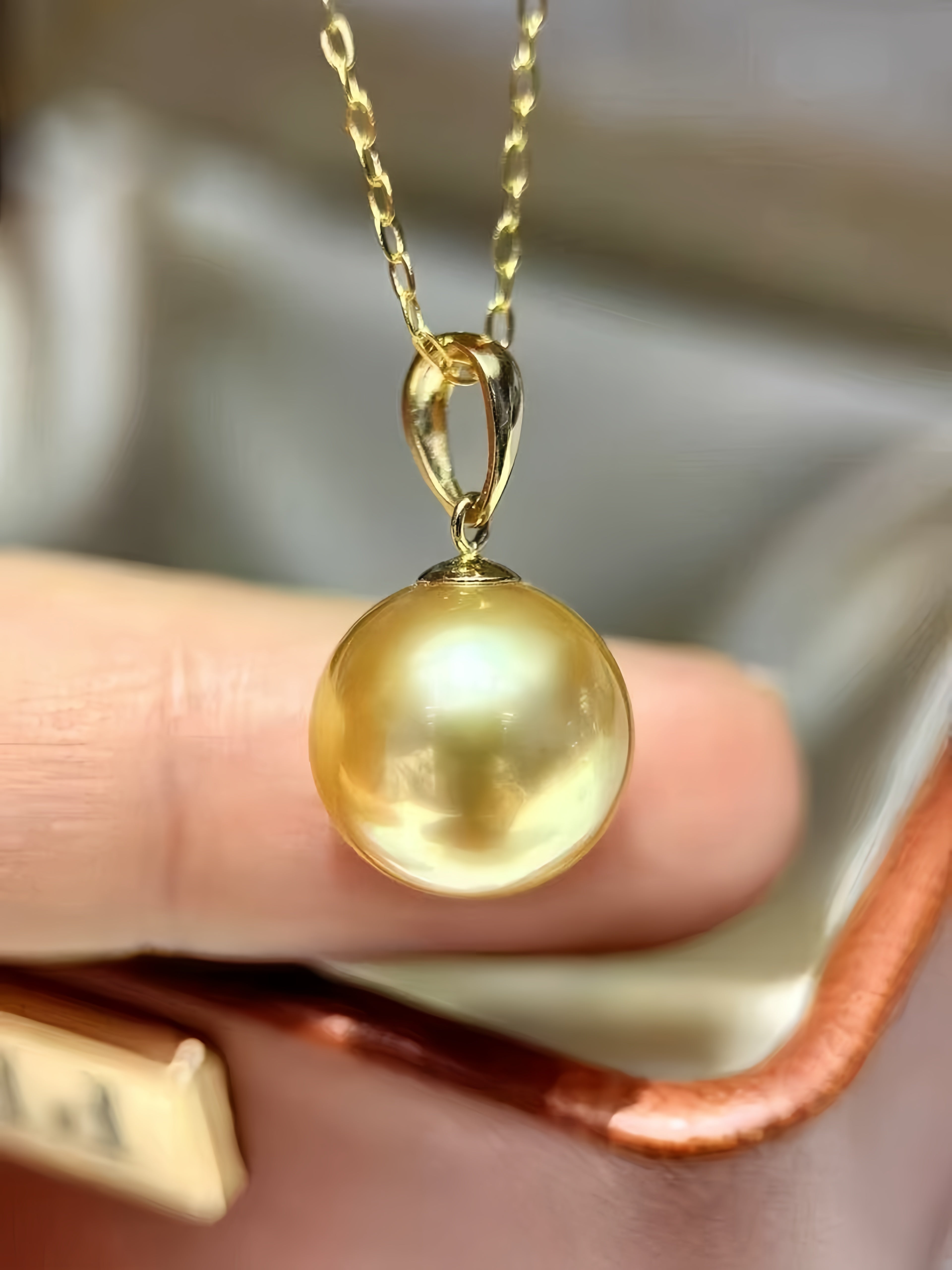 A close-up of a gold pendant necklace featuring a round, lustrous gold pearl on a delicate gold chain, held between fingers.