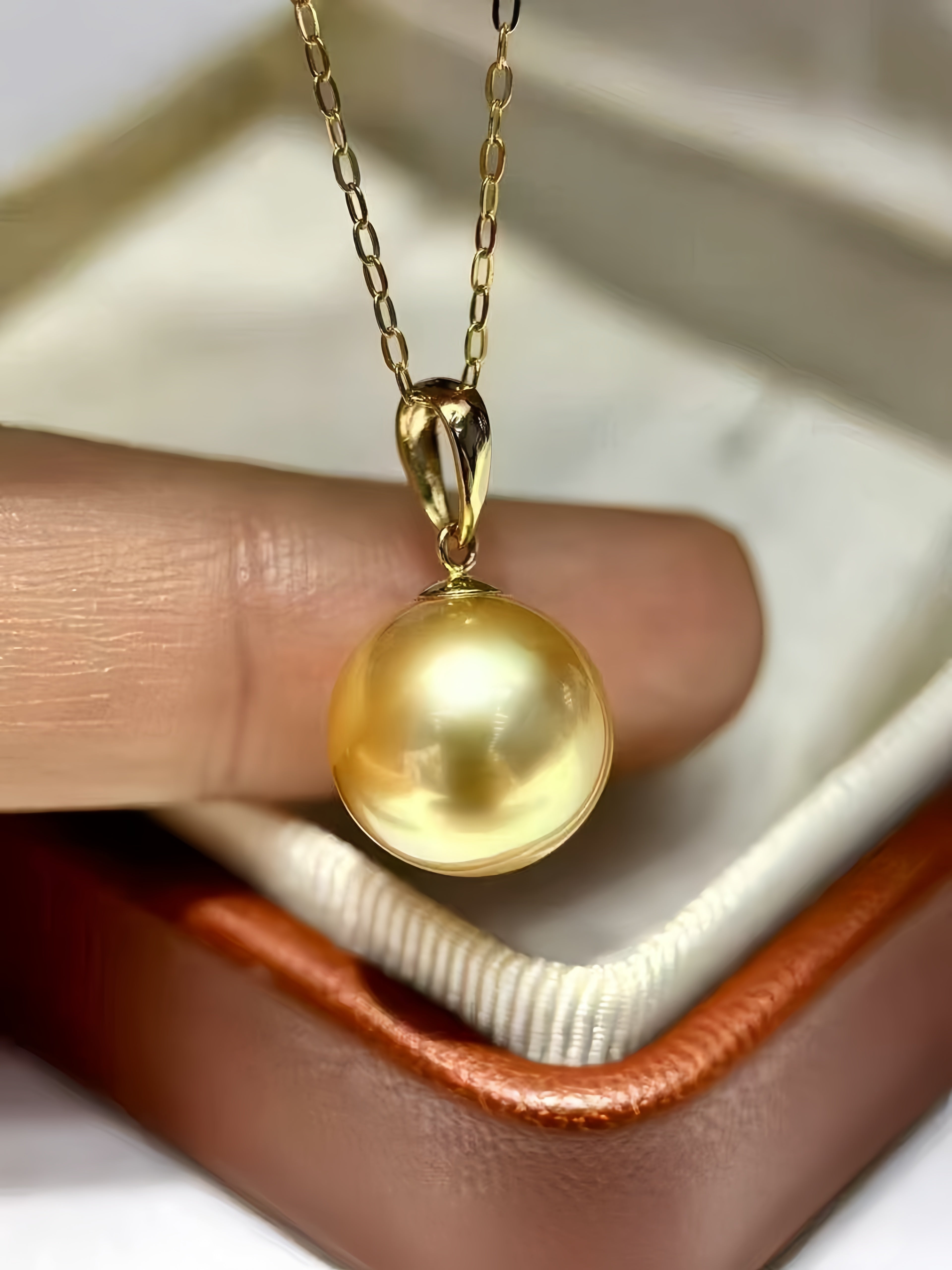 A close-up of a gold pendant necklace featuring a lustrous round pearl-like drop, held by a finger over an open jewelry box.