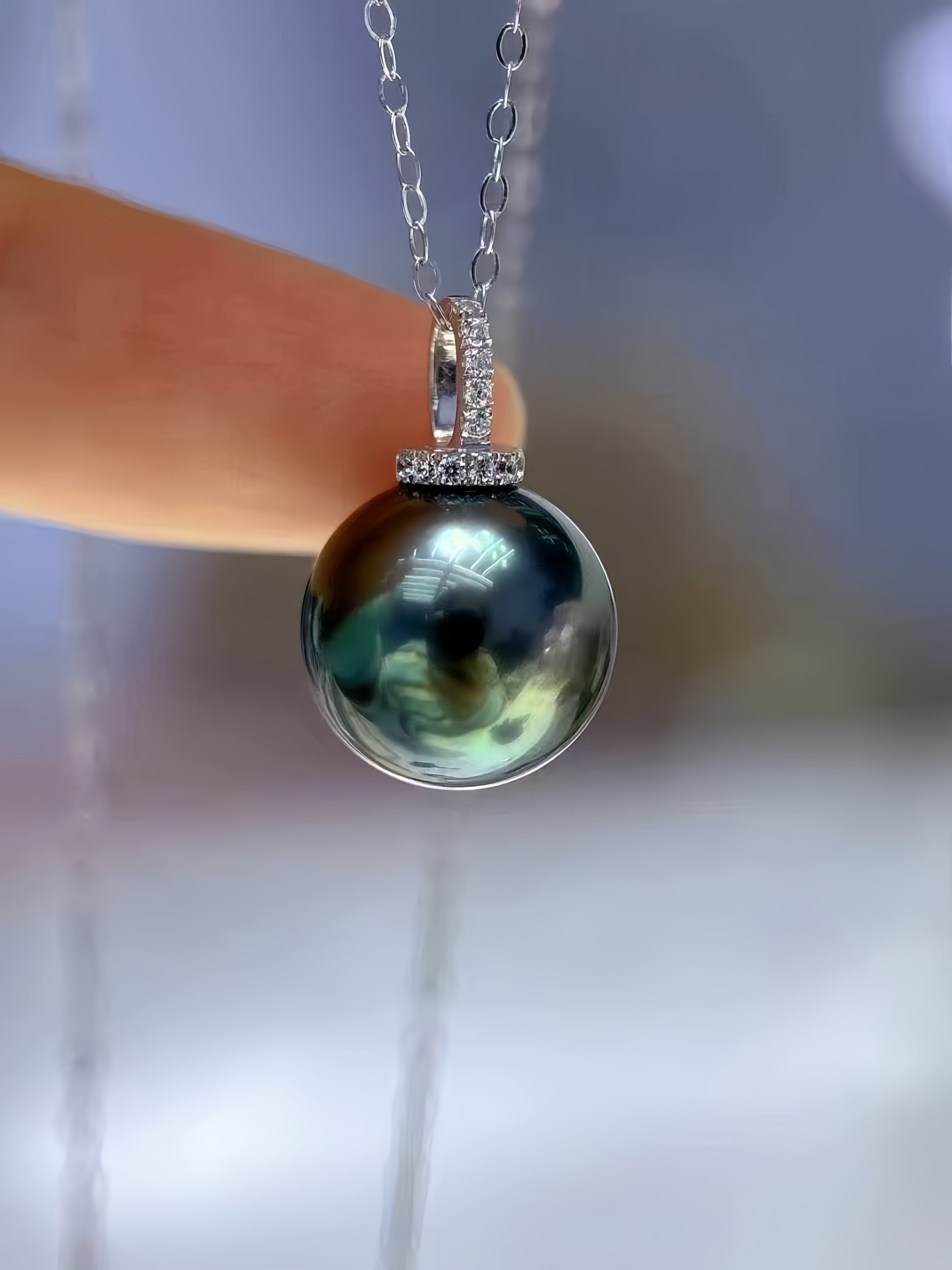 A close-up of a silver pendant necklace held by a finger, featuring a large iridescent dark green pearl pendant with a silver bezel set with small clear stones.