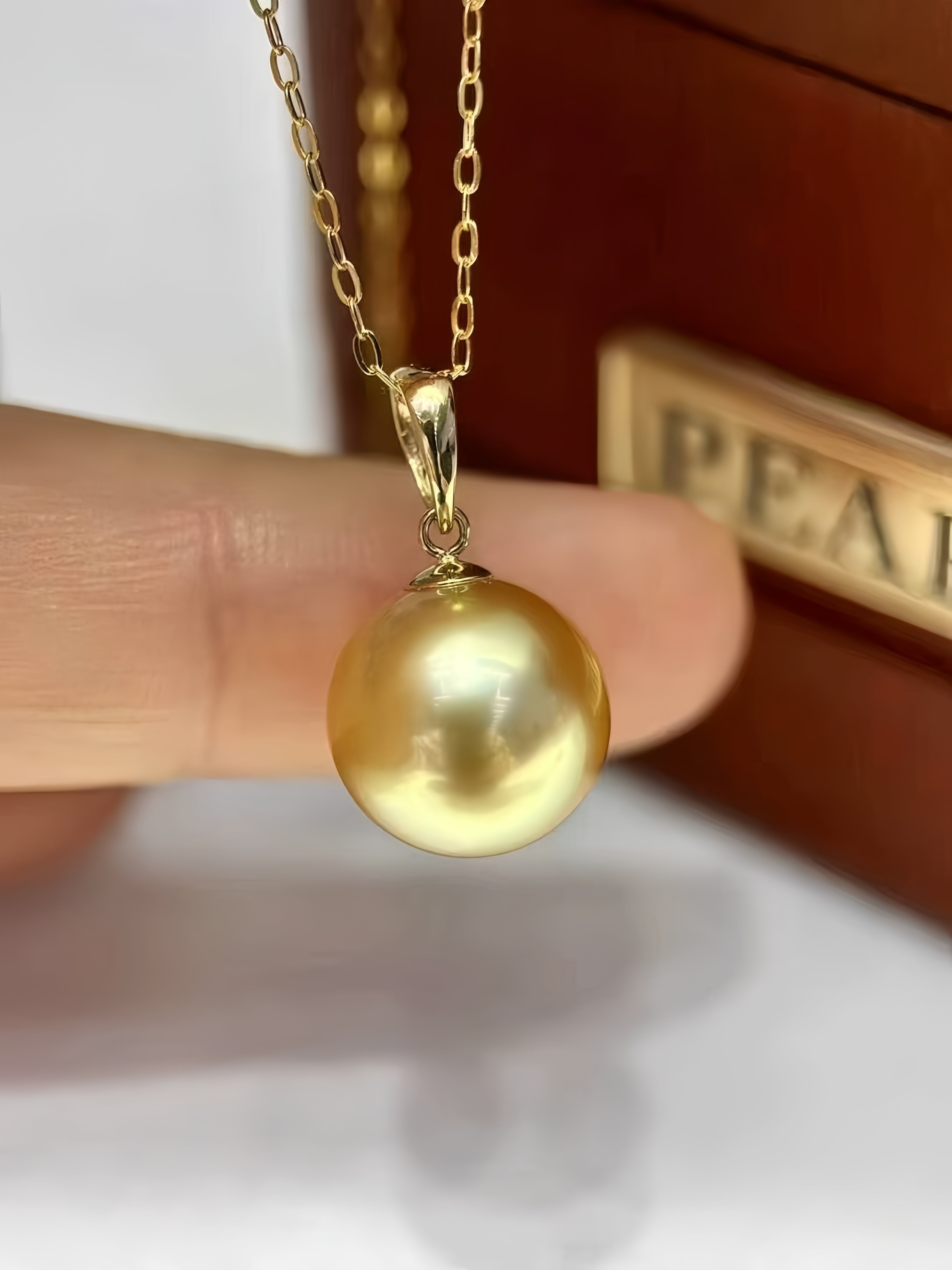 A gold pendant necklace featuring a large, lustrous round pearl, held by a person's fingers in front of a brown jewelry box.