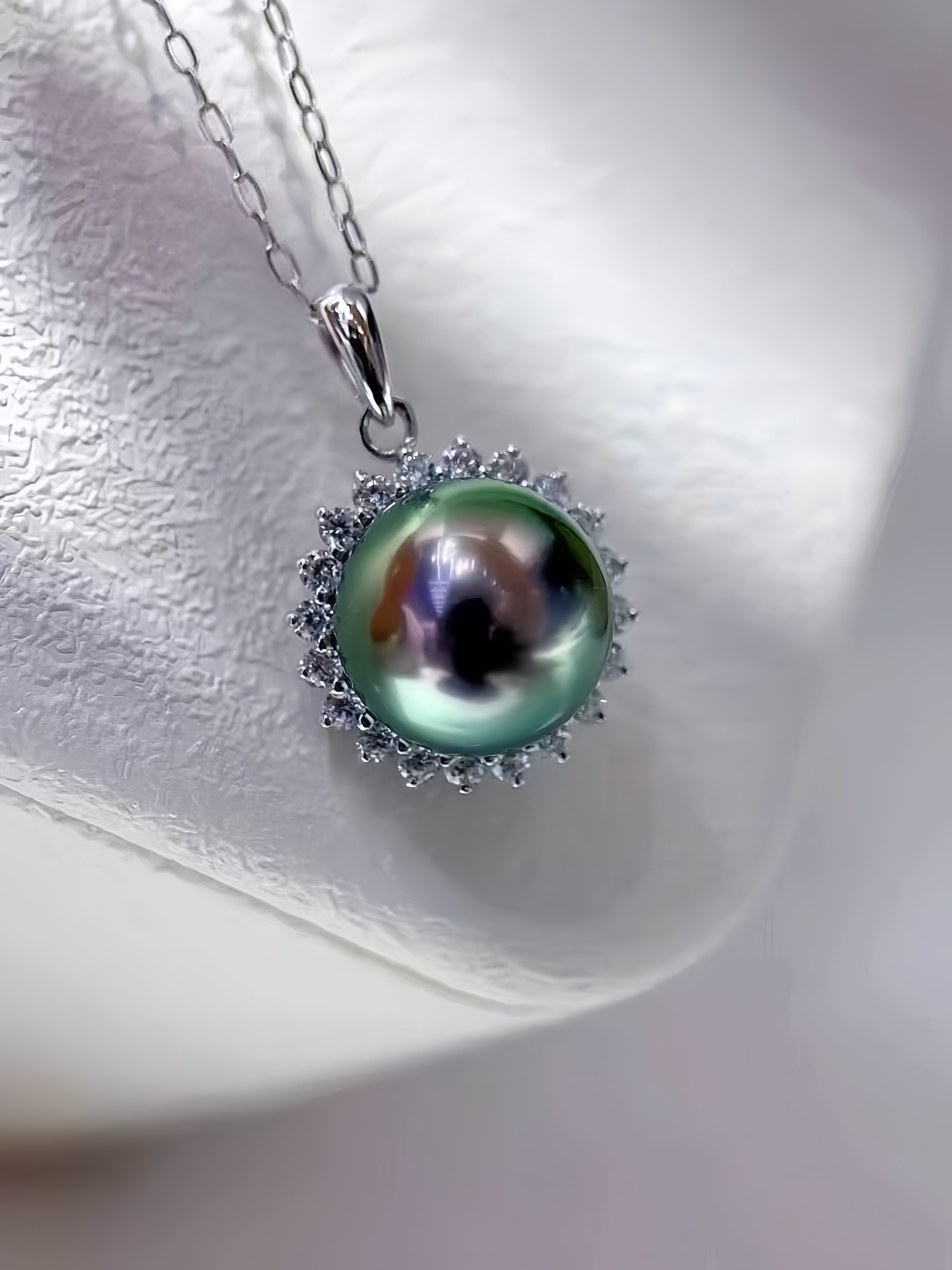 A silver pendant necklace featuring a large, iridescent green pearl surrounded by a halo of small clear stones, resting on a textured white display stand.