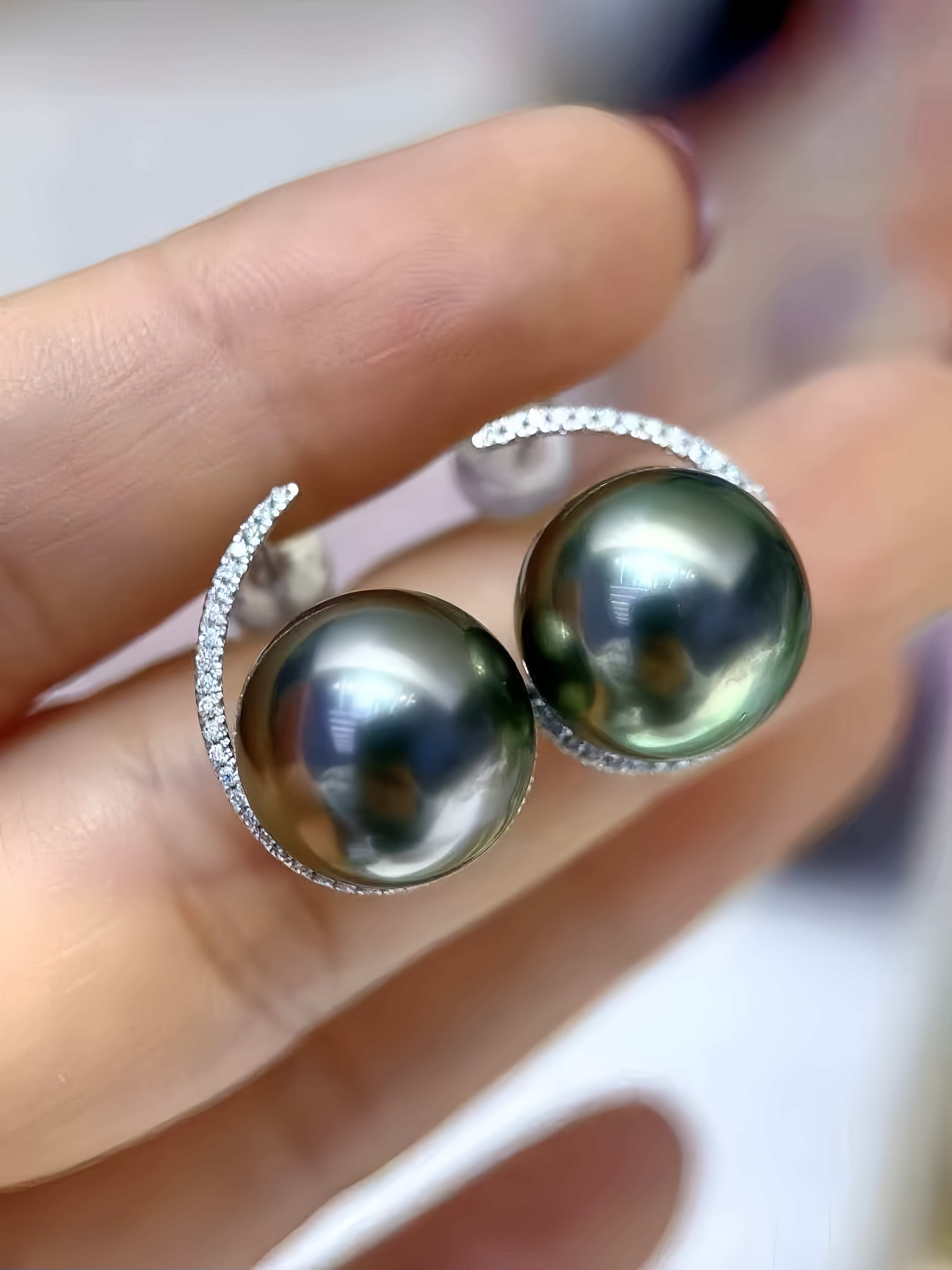 A pair of silver crescent-shaped earrings with round dark green-black Tahitian pearls and pavé-set clear stones, shown worn on a person's finger.