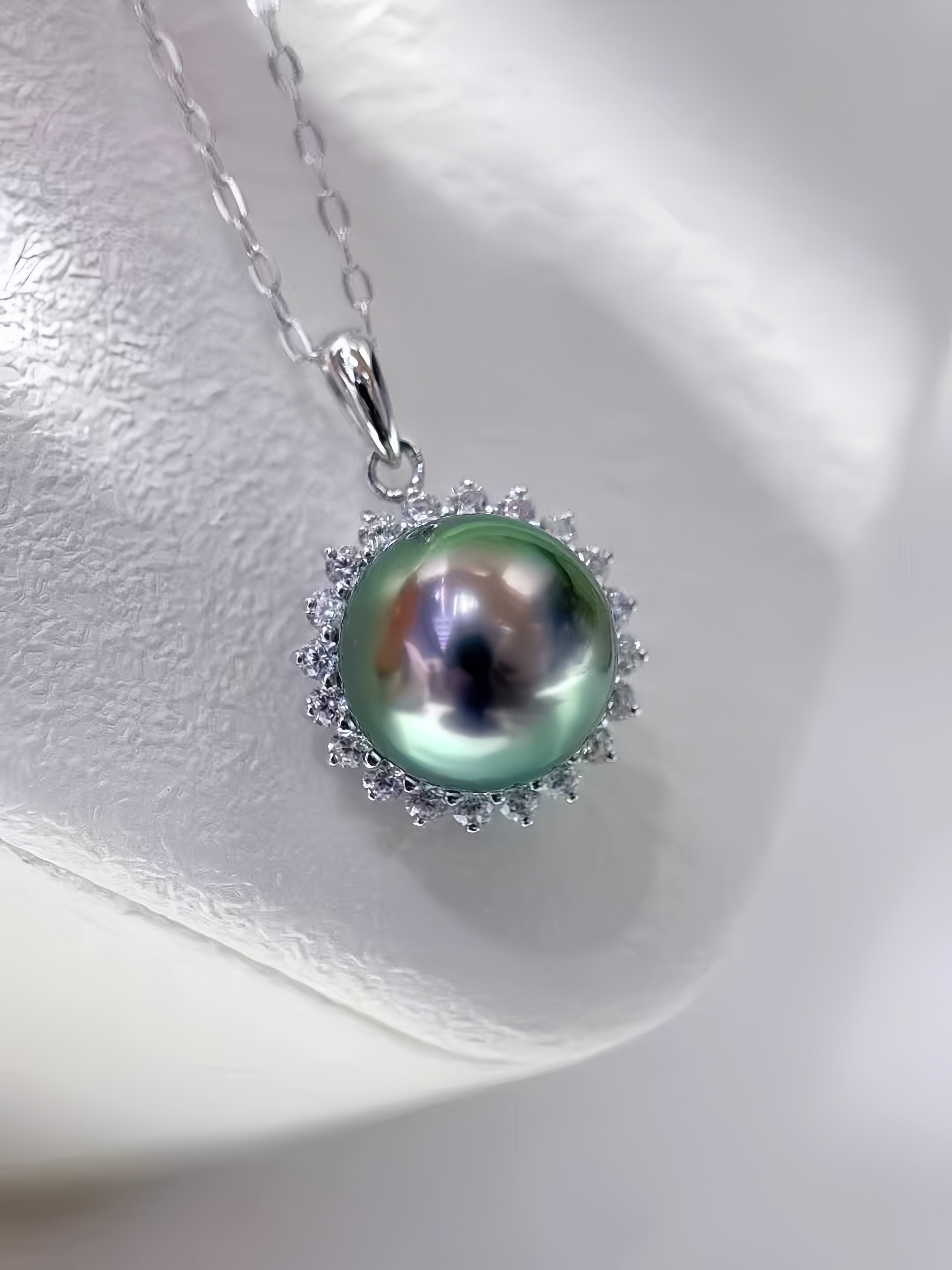 A silver pendant necklace featuring a large, lustrous green pearl surrounded by small clear stones, displayed on a textured white surface.