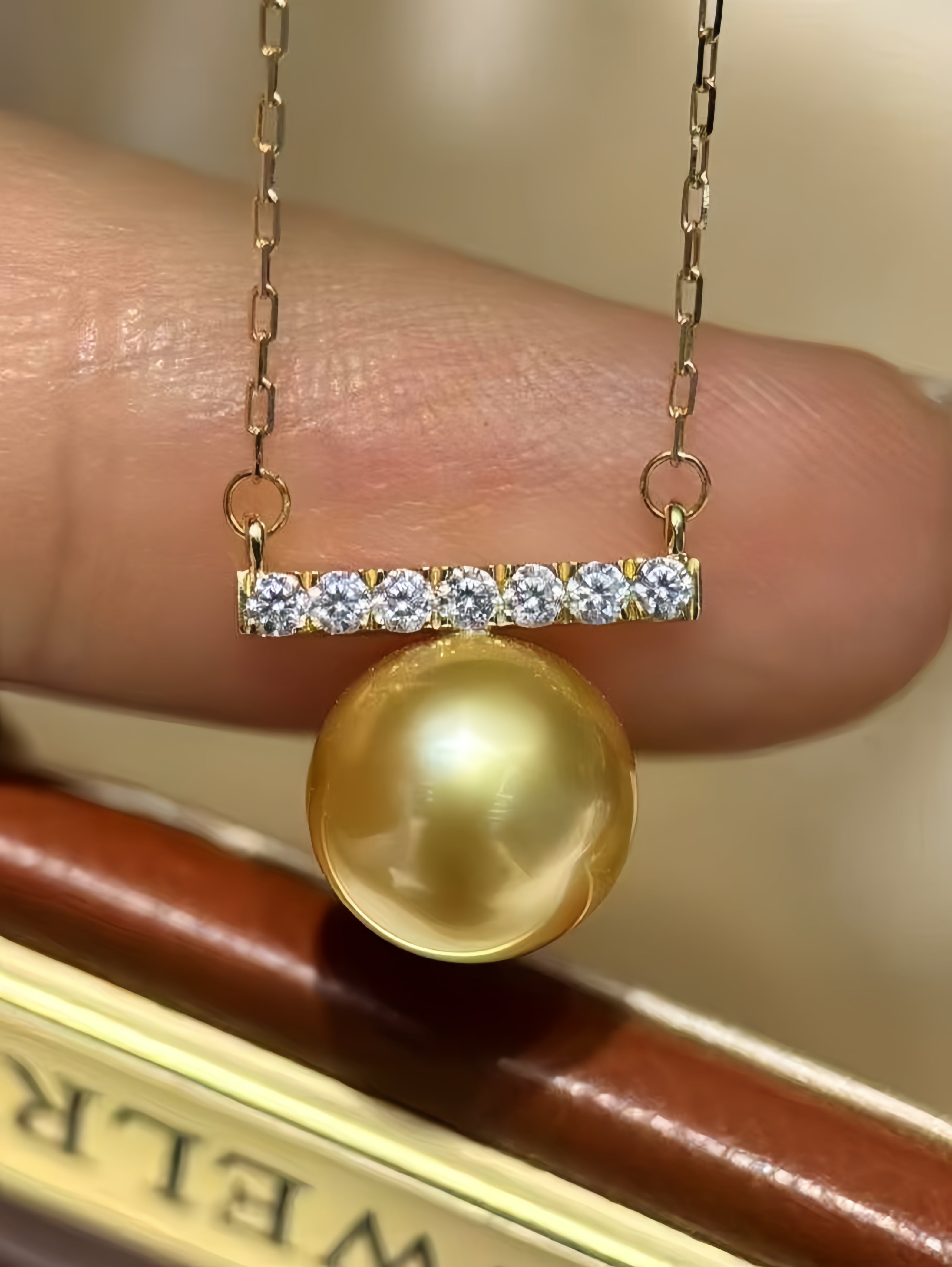 A gold necklace with a bar set with clear stones and a large round gold pearl pendant, held against a person's finger.