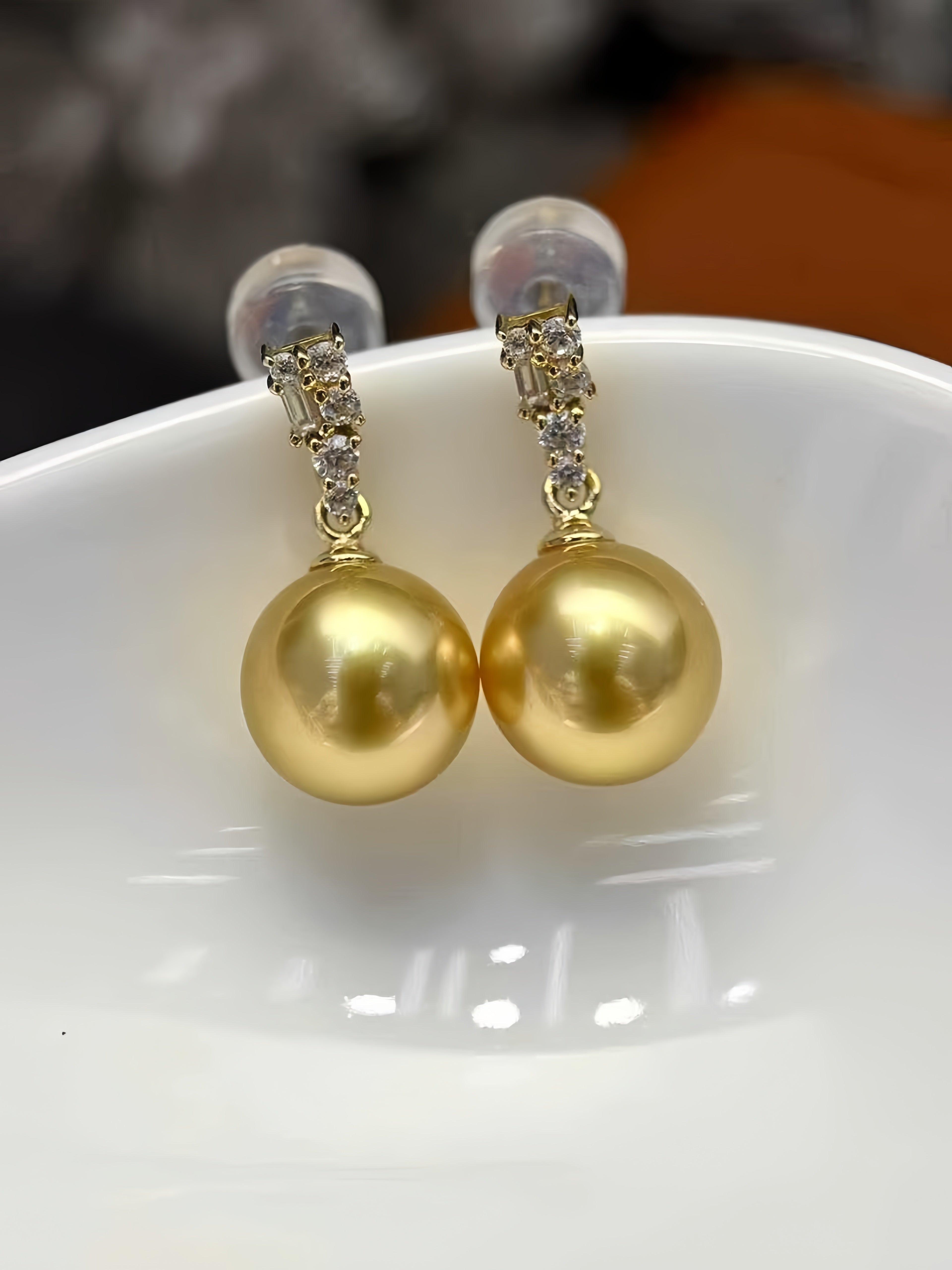 A pair of gold drop earrings with large round gold pearls and small clear stone accents at the top, displayed on a white ceramic dish.