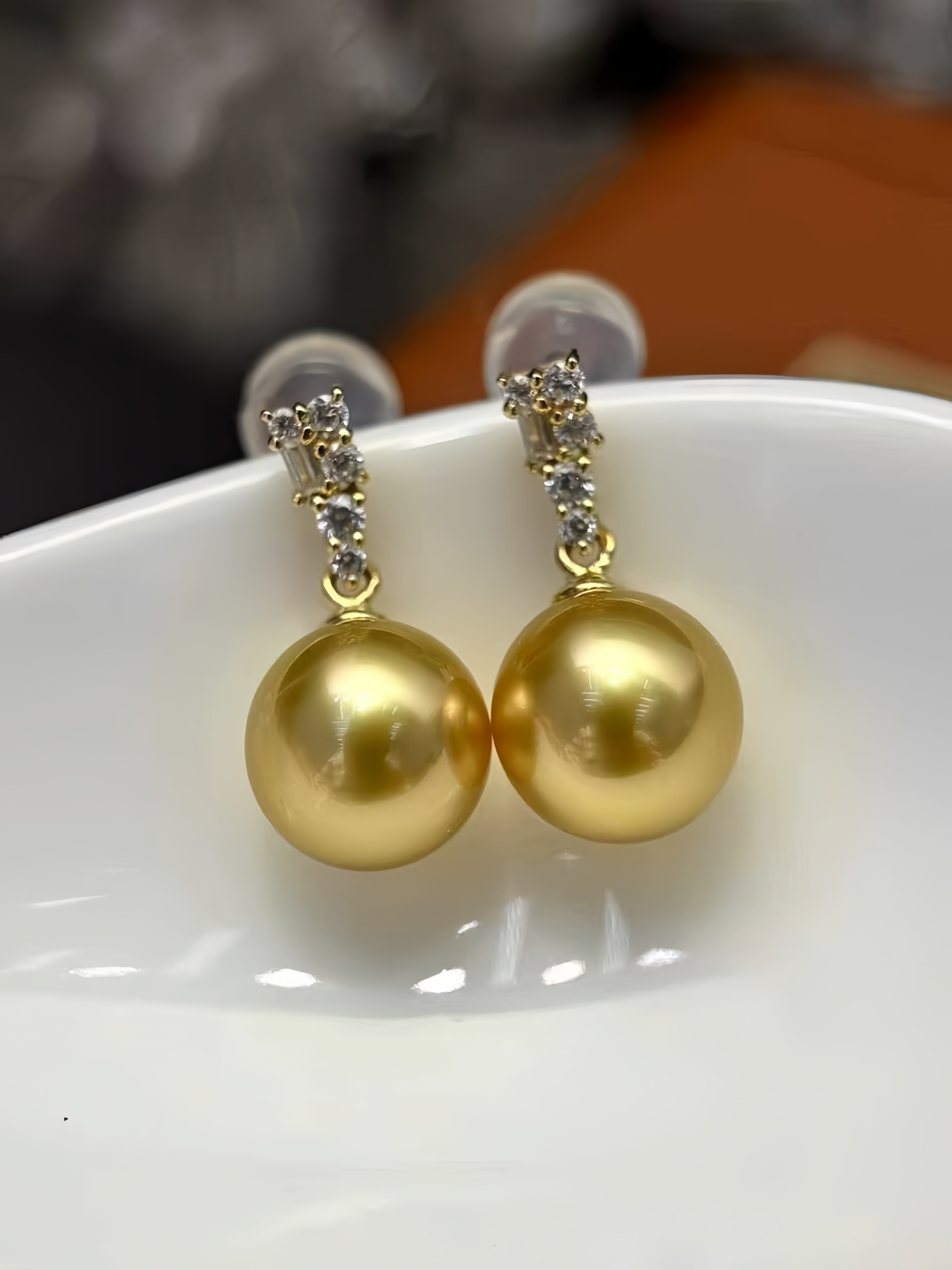 A pair of gold-toned drop earrings with a small cluster of clear stones above a large round gold pearl, displayed on a white ceramic dish.