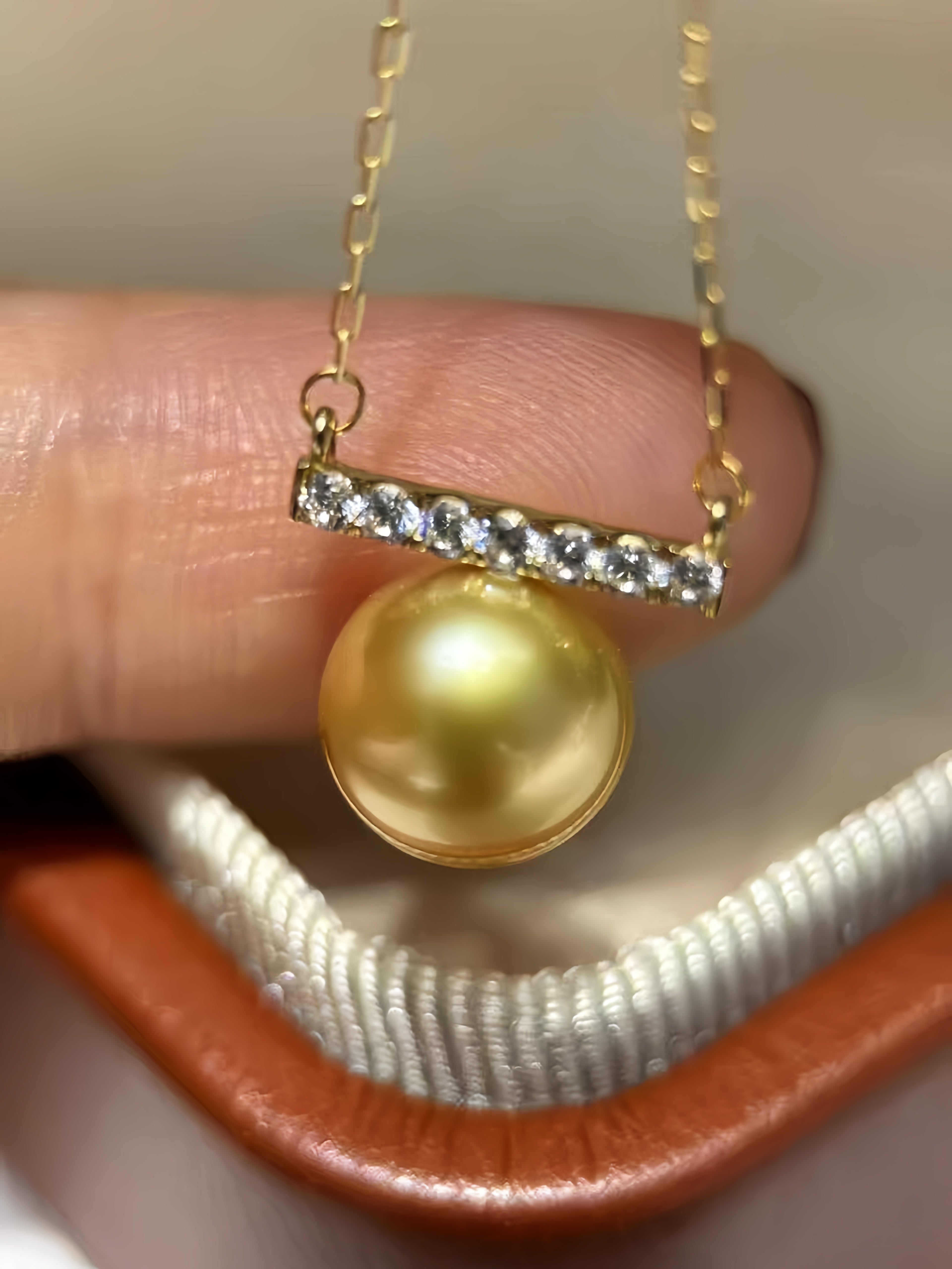 A gold pendant necklace featuring a round yellow pearl and a bar-set clear crystal accent, held between fingers over a white and brown jewelry box.