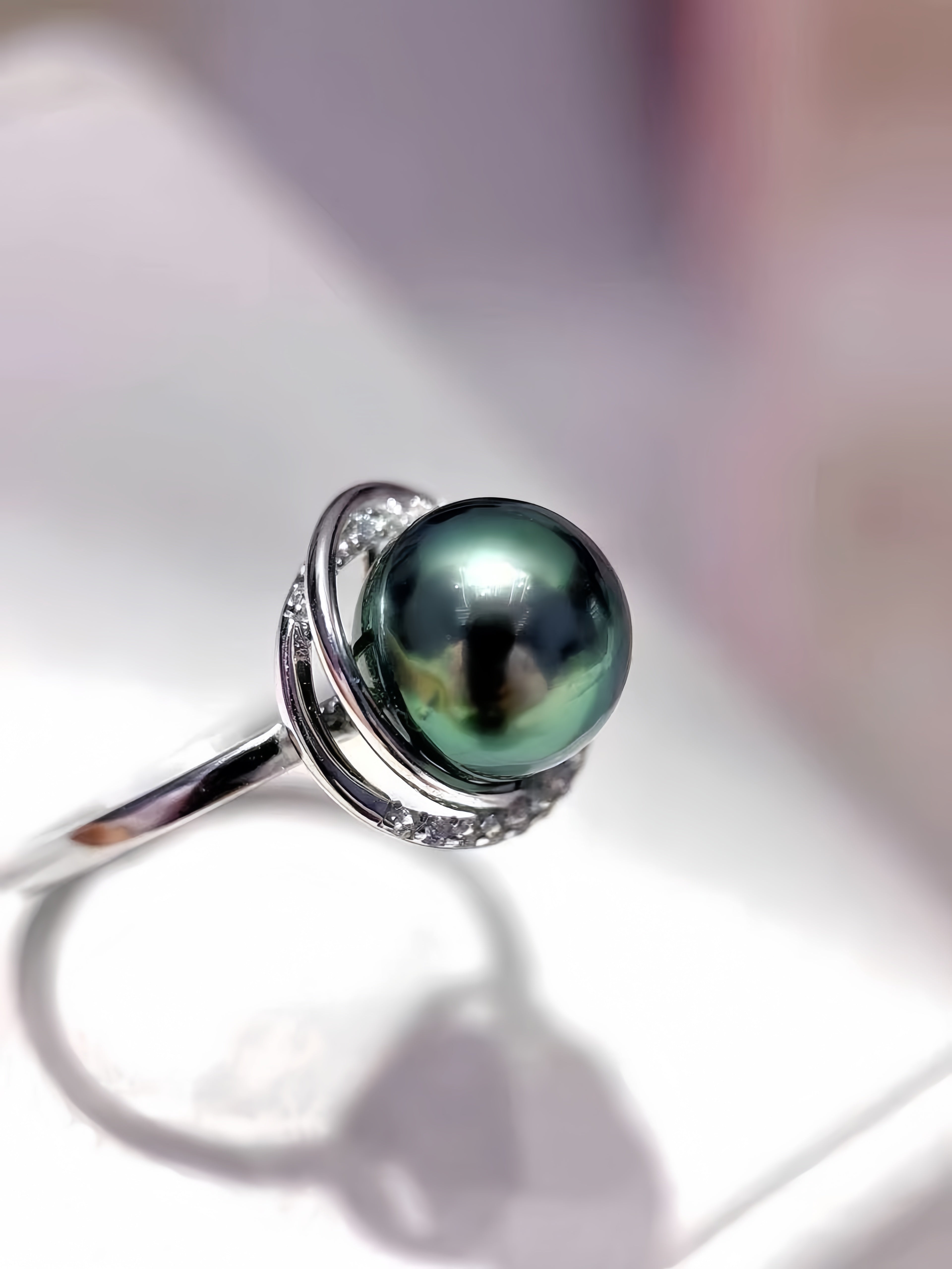 A sterling silver ring featuring a large, round, iridescent green-black Tahitian pearl set in a double-ring halo with small accent stones, shown on a soft-focus white background.