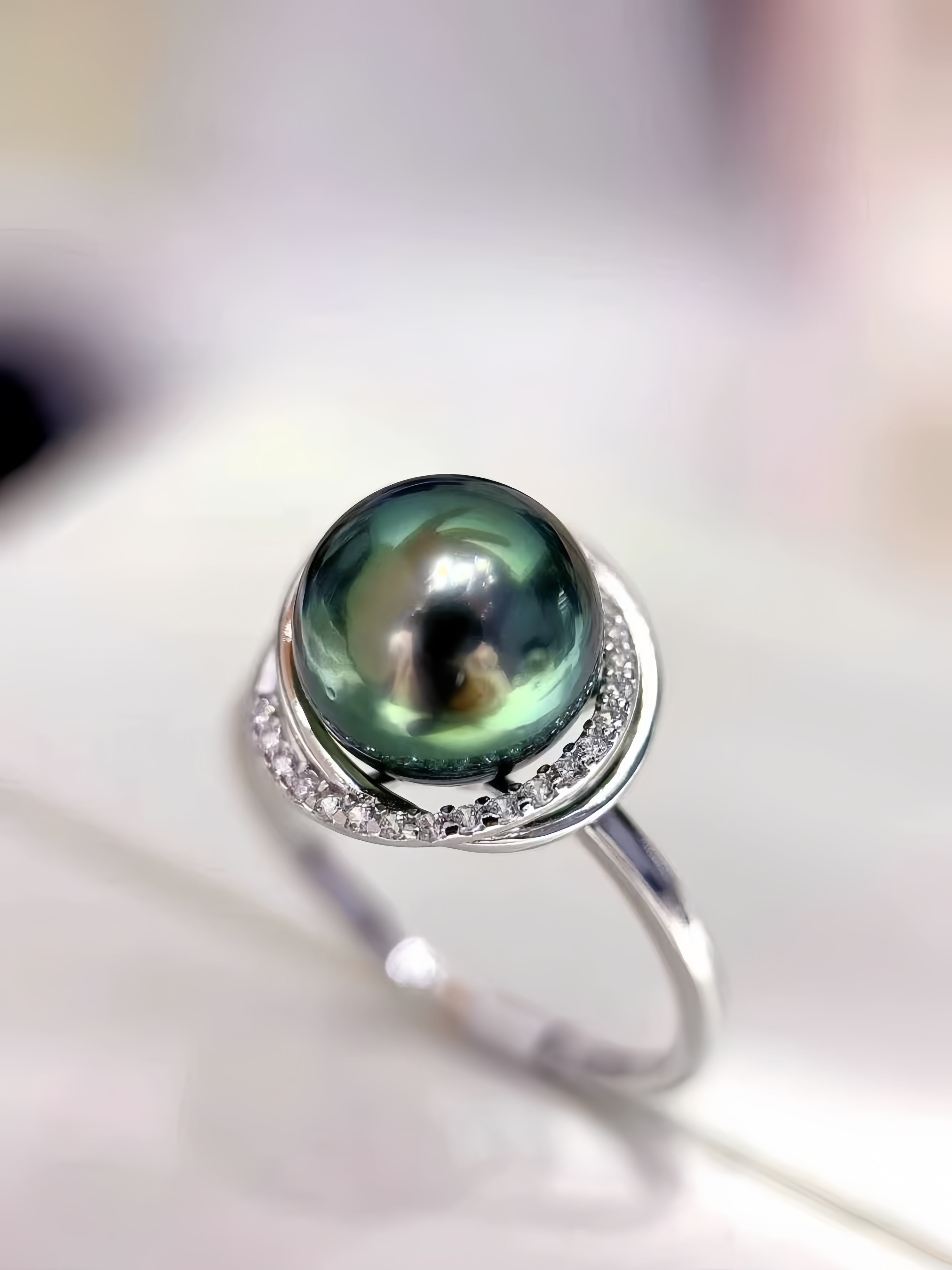 A sterling silver ring featuring a large, round green-black Tahitian pearl in a bezel setting with a halo of small clear stones, shown on a soft-focus light background.