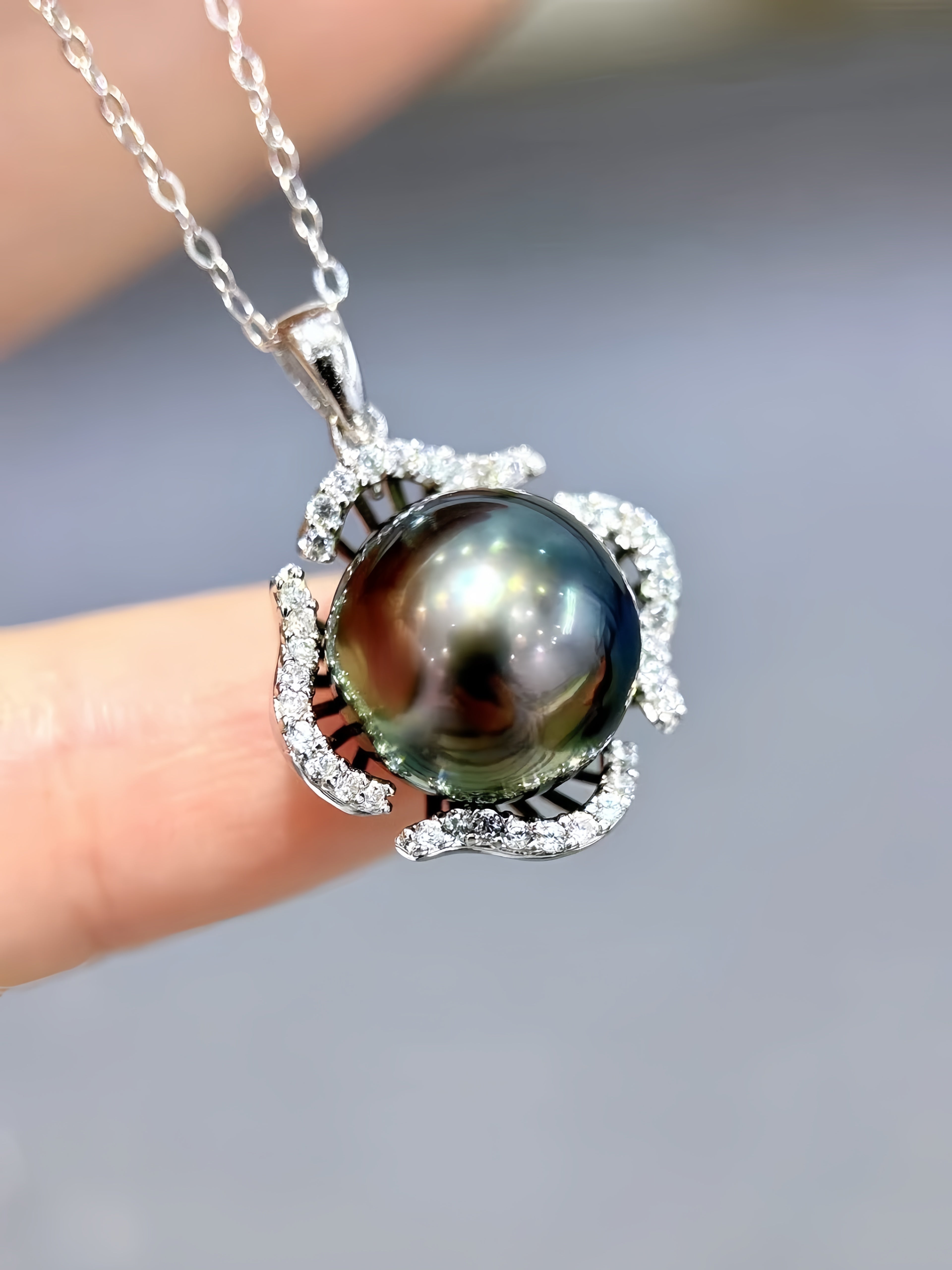 A close-up of a silver pendant necklace featuring a large, round, dark green-black Tahitian pearl surrounded by small clear stones, held by a person's fingers against a soft gray background.