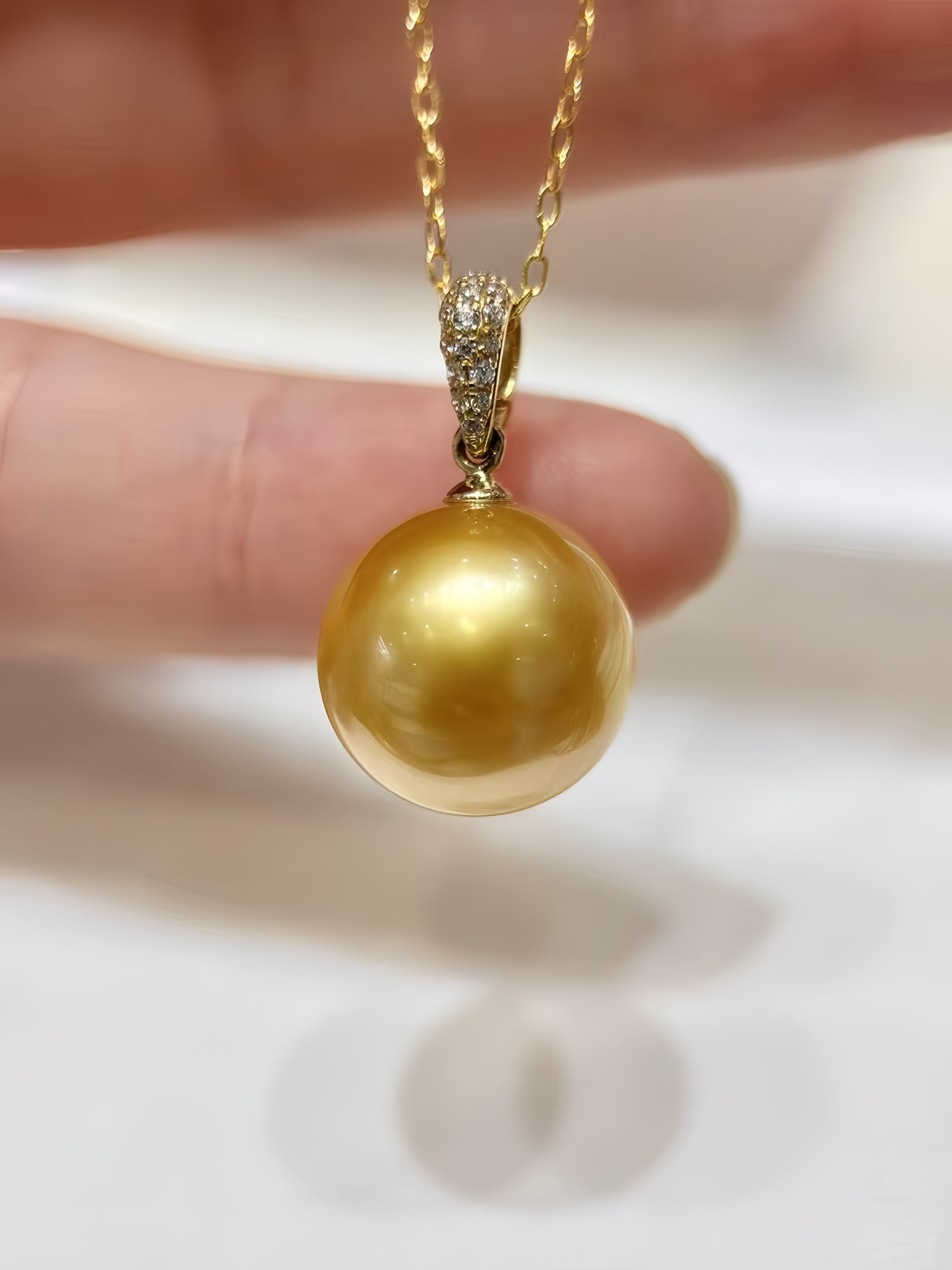 A close-up of a gold pendant necklace featuring a large, lustrous golden pearl pendant and a gold chain, held by fingers against a soft white background.