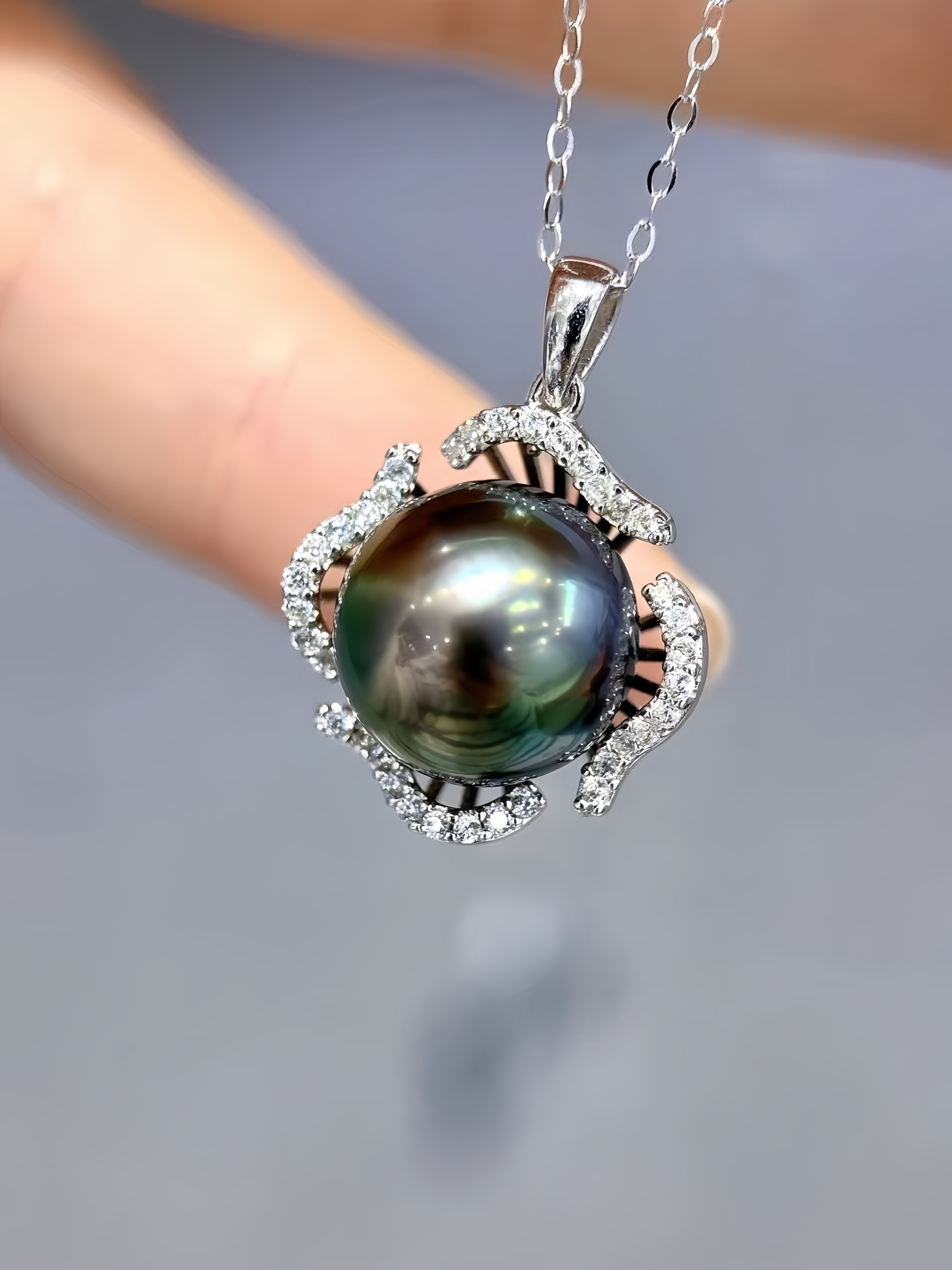 A close-up of a silver pendant necklace featuring a large, iridescent green-black freshwater pearl surrounded by small clear stones, held by a person's fingers against a soft gray background.