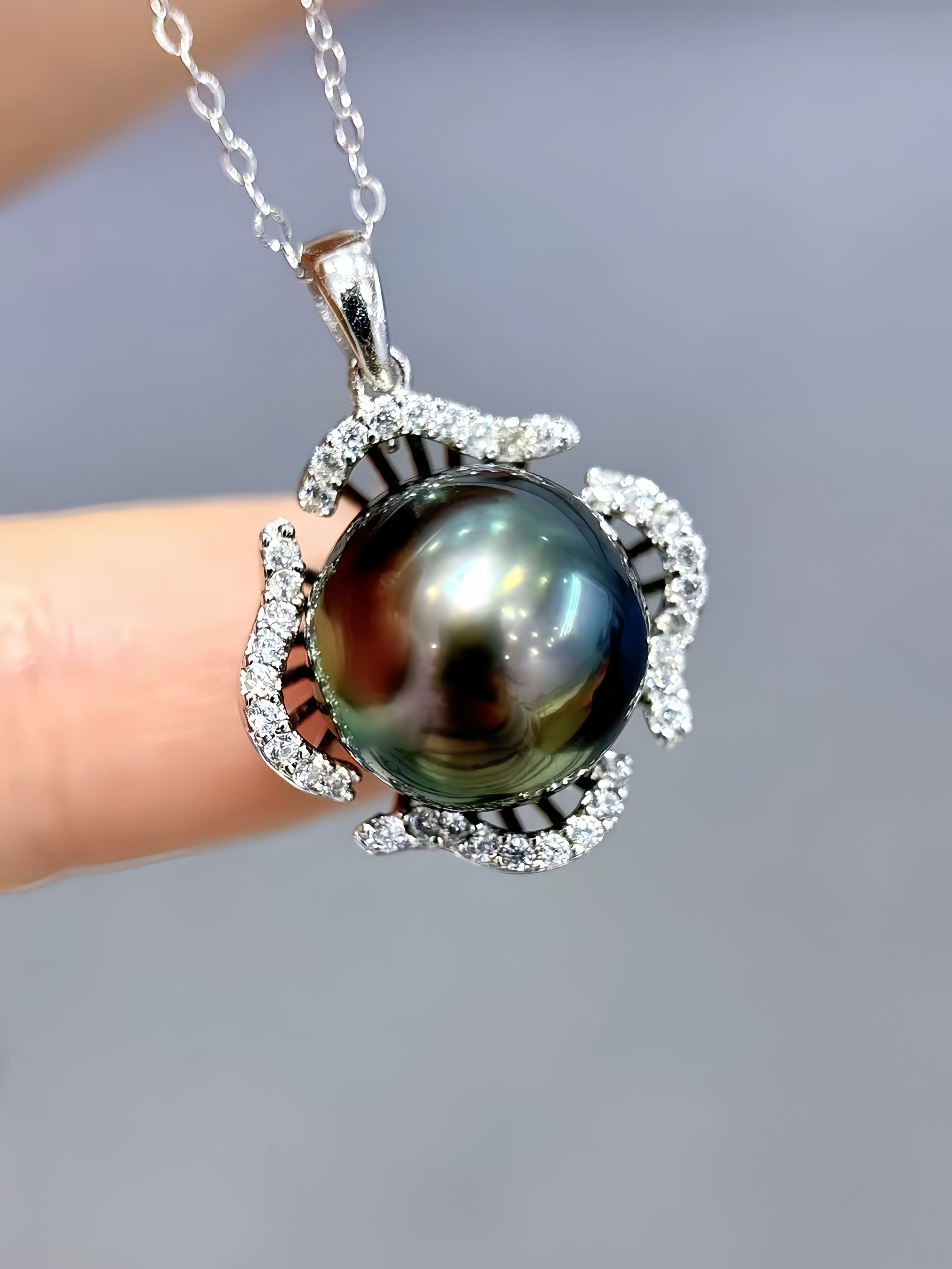 A close-up of a silver pendant necklace featuring a large, iridescent dark green pearl surrounded by small clear stones, held between fingers against a soft gray background.