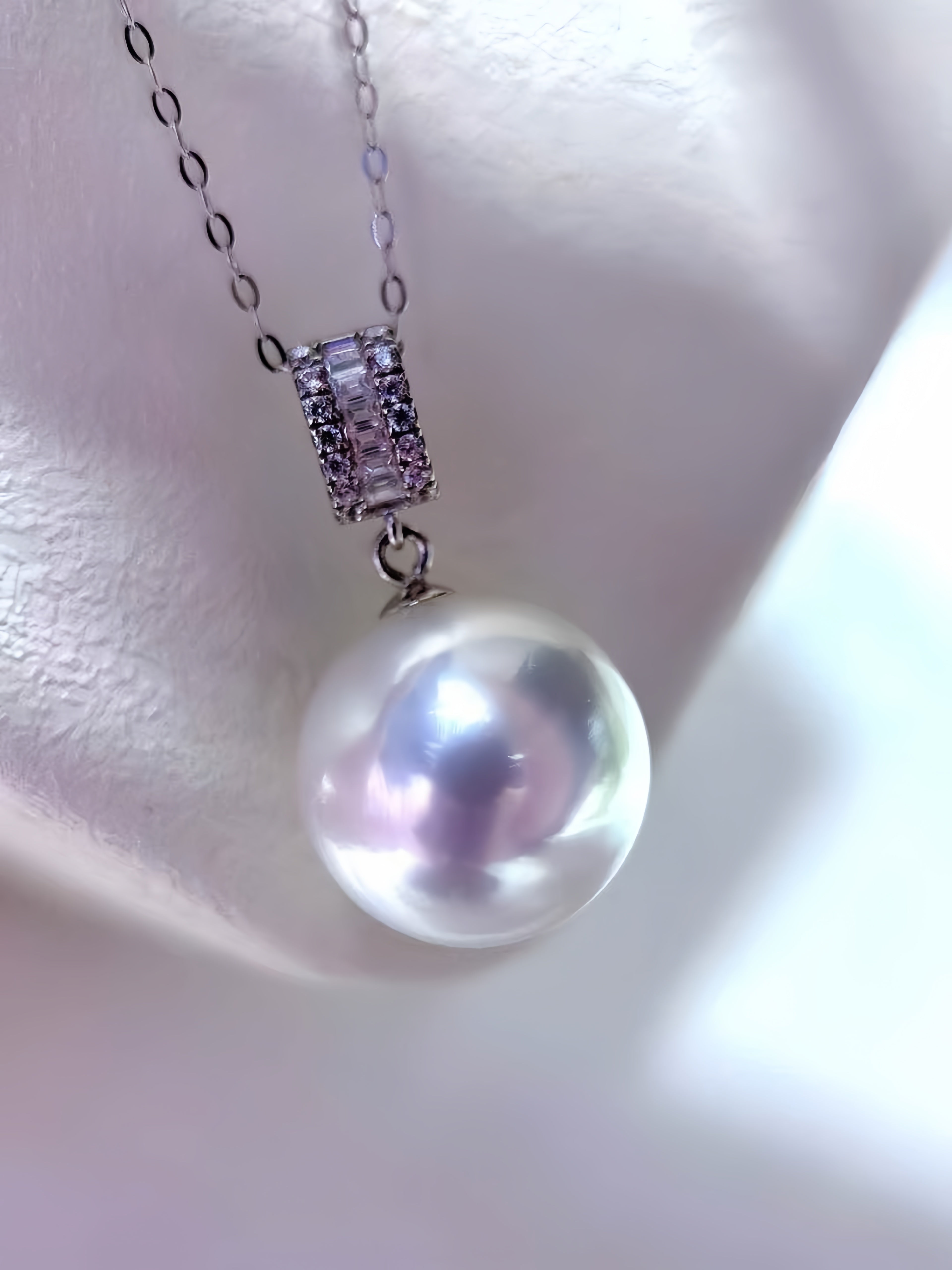 A close-up of a pearl pendant necklace with a silver-toned chain and a rectangular pavé-set bezel containing clear stones, displayed on a white jewelry bust.