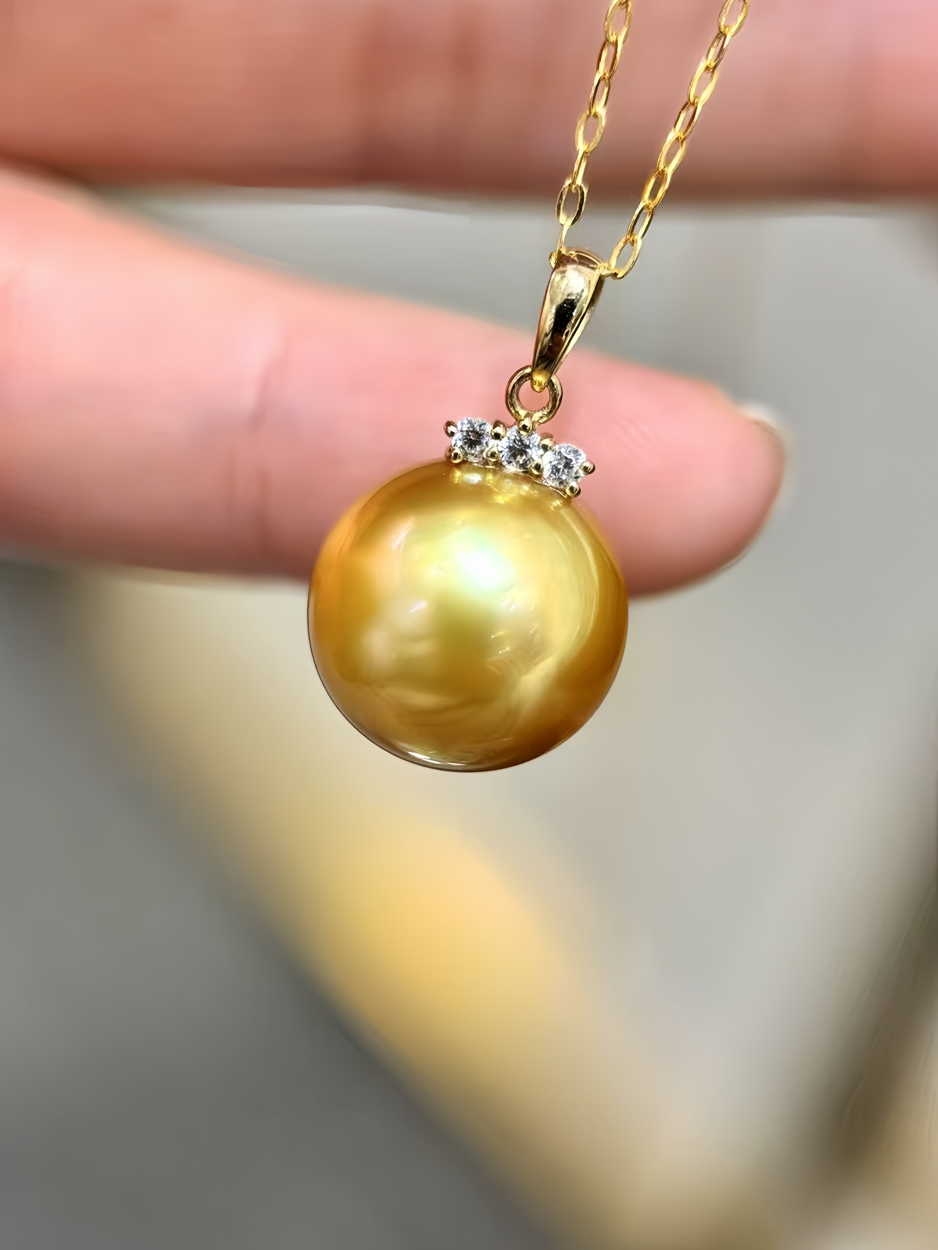 A close-up of a gold pendant necklace featuring a large, lustrous golden pearl pendant with a small cluster of clear stones at the top, held by fingers.