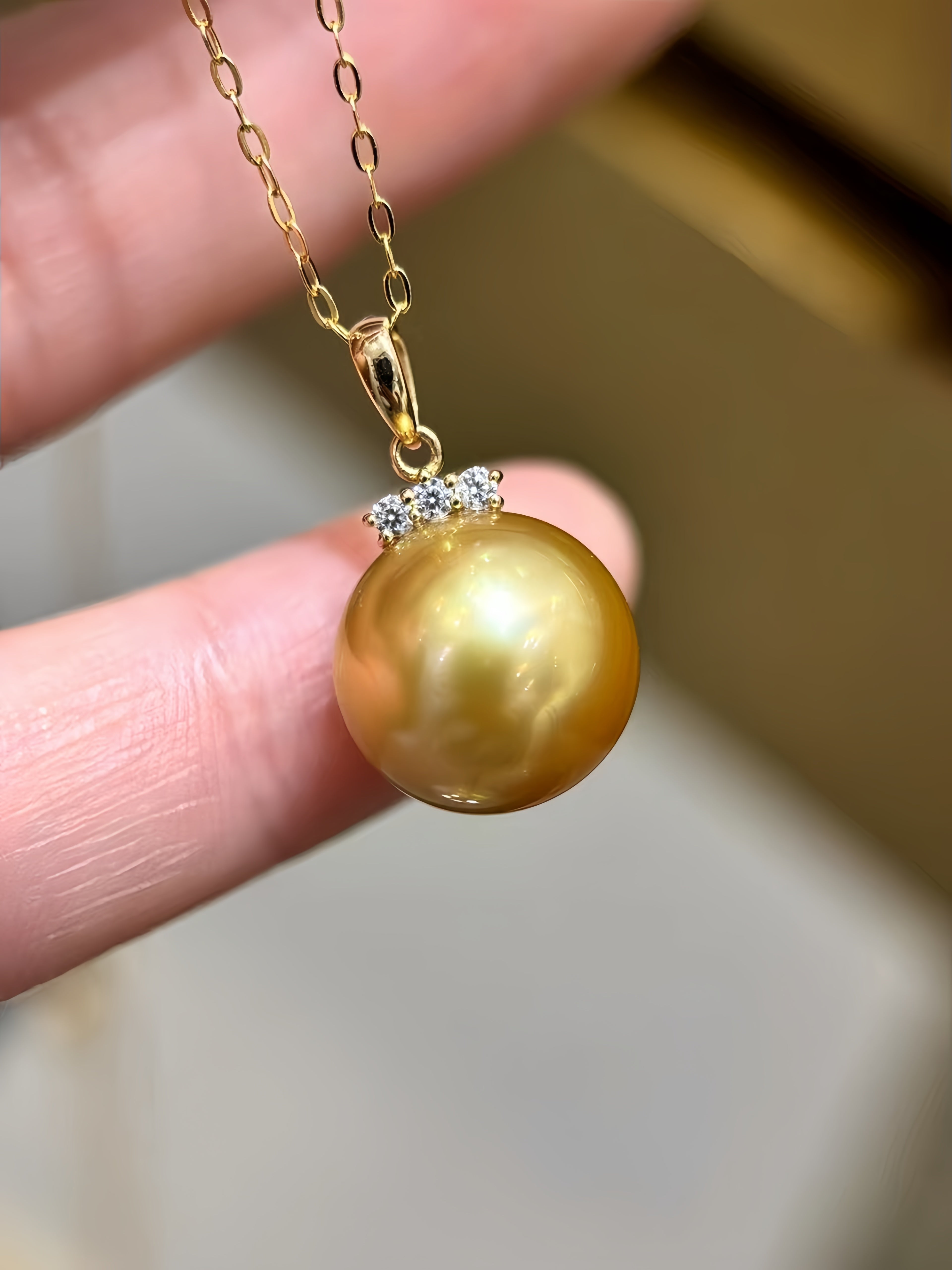A gold pendant necklace featuring a large, round, lustrous gold pearl accented by a small cluster of clear stones at the top, held by a person's fingers against a soft-focus background.
