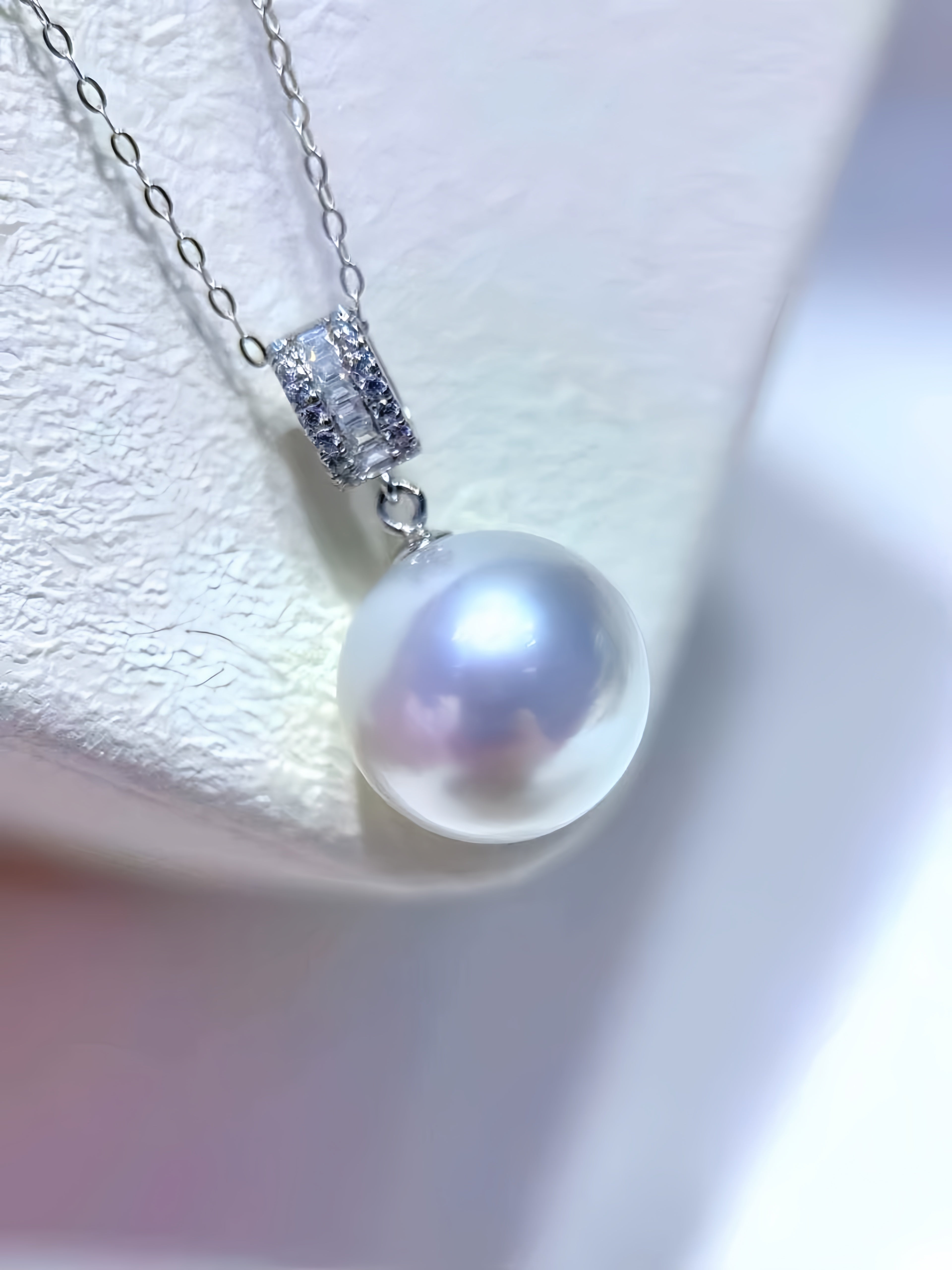 A close-up of a silver chain necklace with a large, lustrous white pearl pendant and a sparkling silver bezel setting, displayed on a textured white jewelry stand.