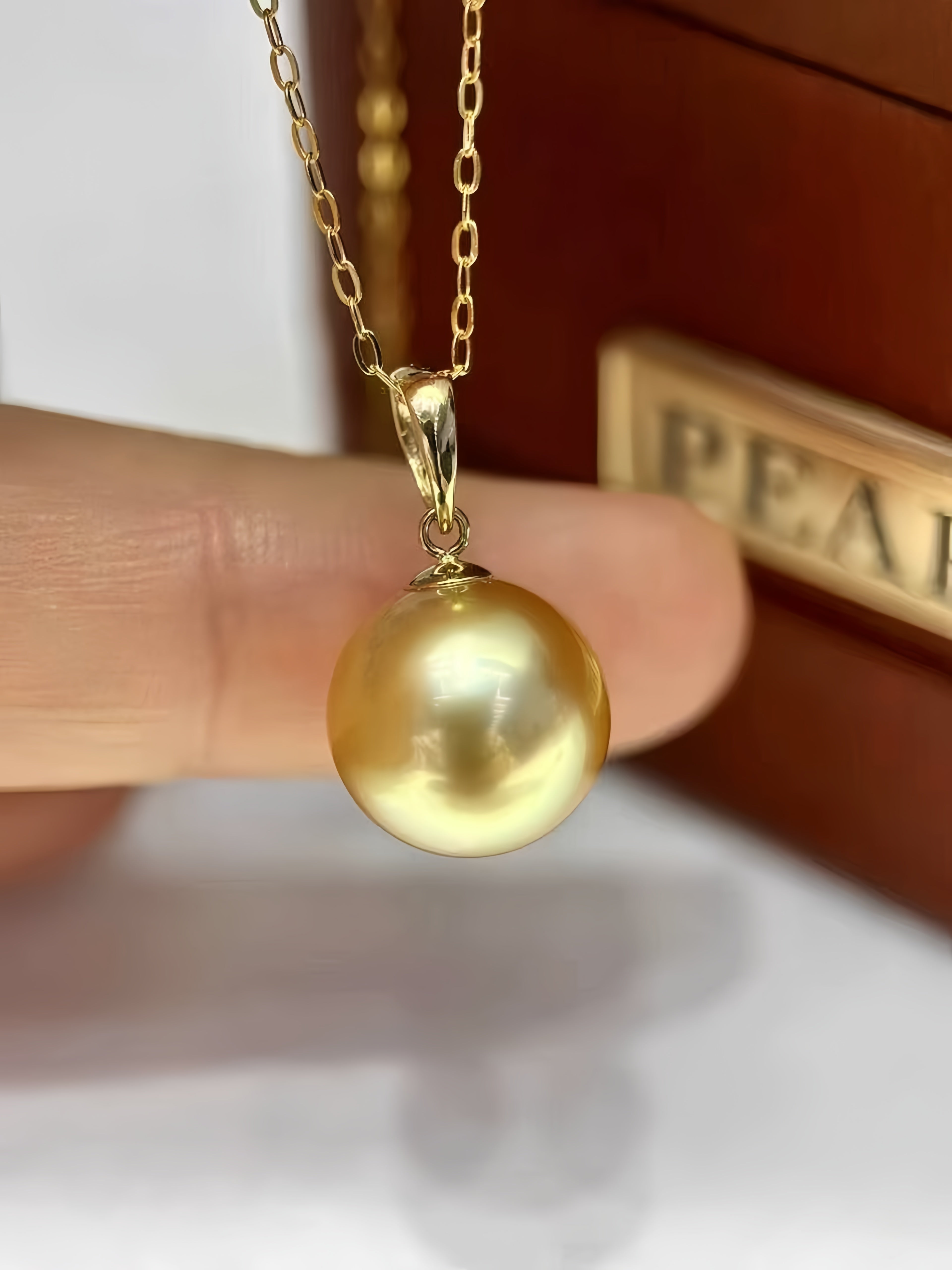 A gold pendant necklace featuring a large, lustrous round pearl, held by a person's fingers in front of a brown jewelry box.