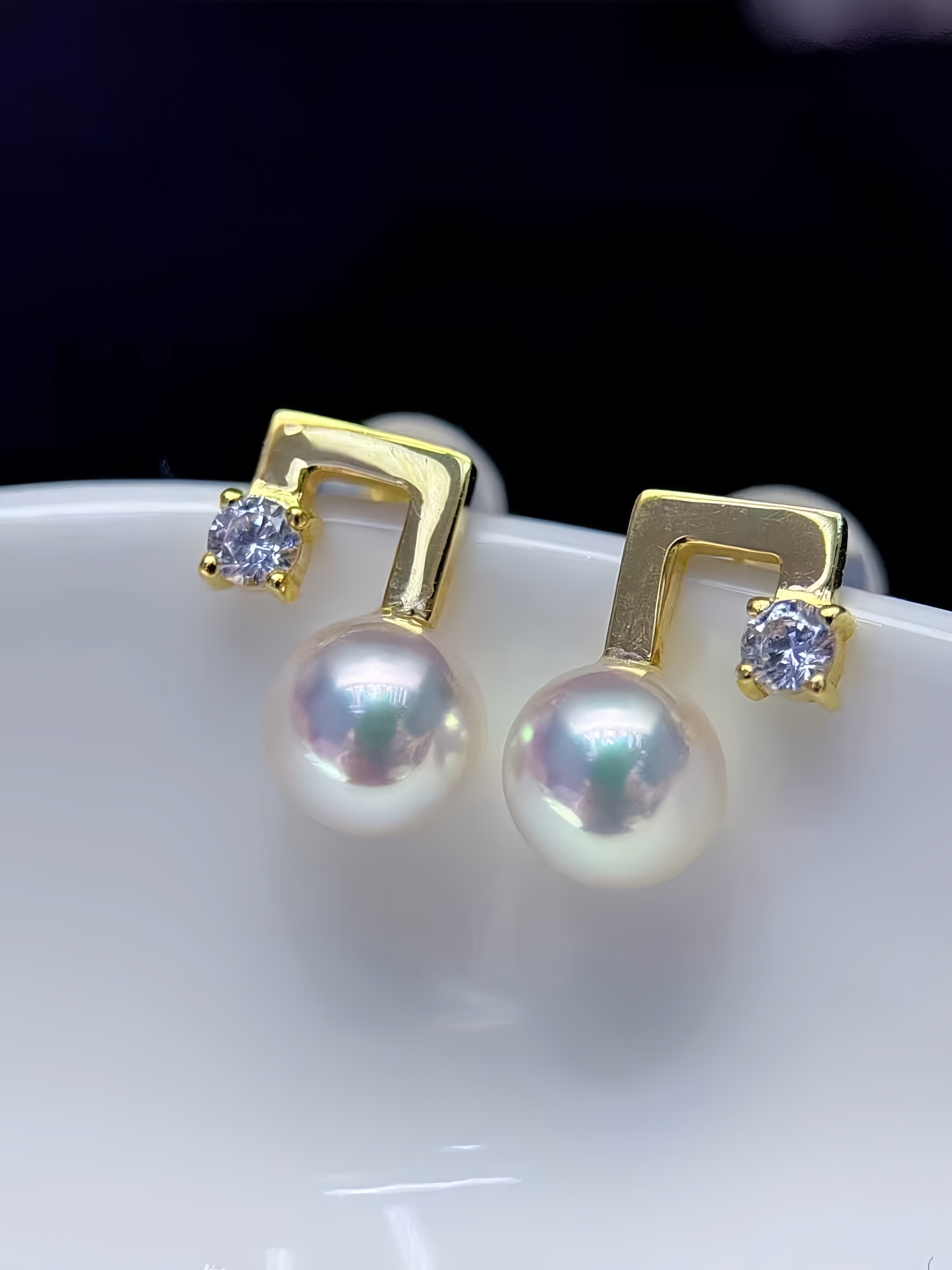 A pair of gold and pearl stud earrings displayed on a white surface against a dark background. Each earring features a gold geometric post with a small clear stone and a large iridescent white pearl hanging below.