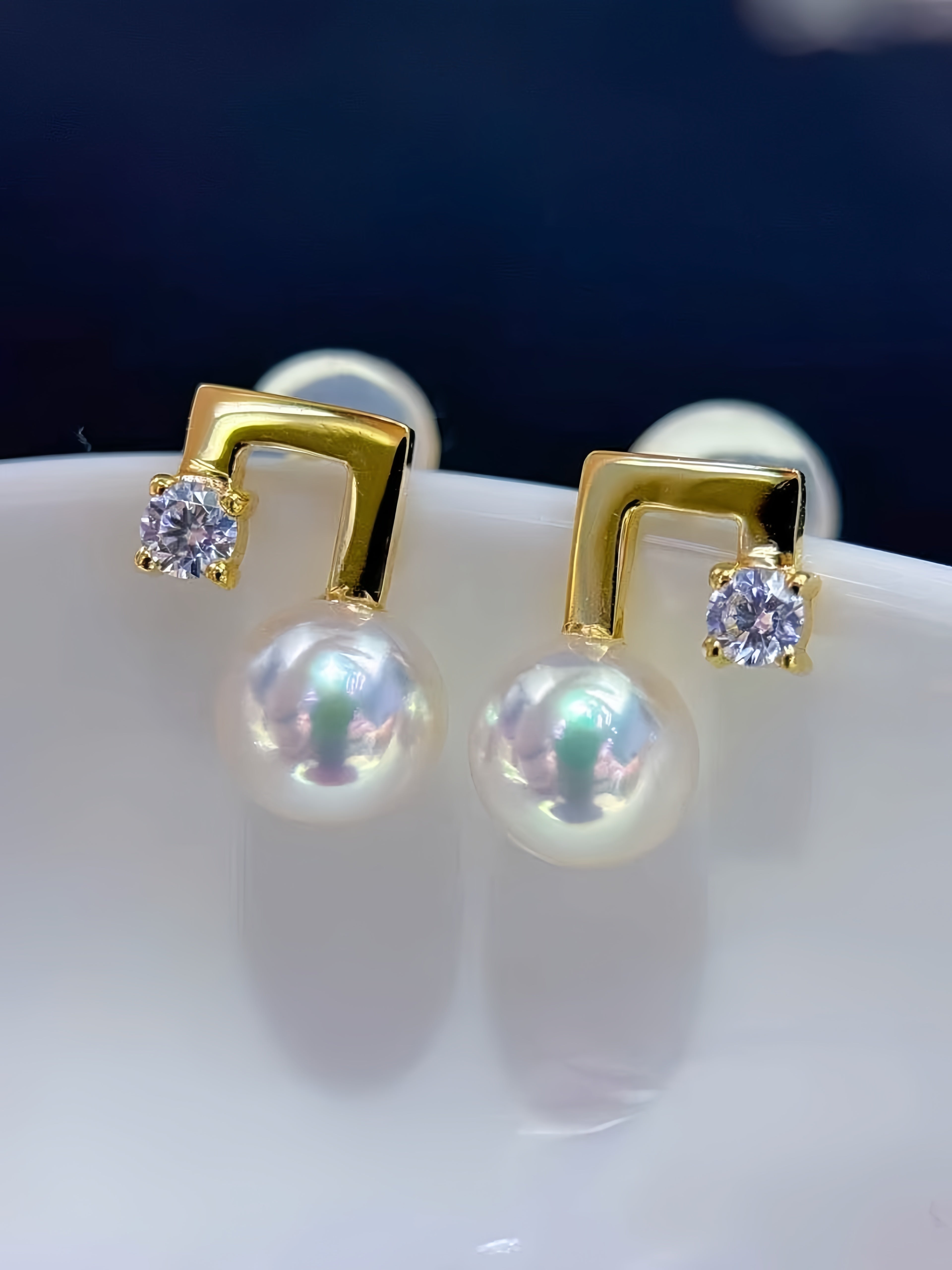 A pair of gold and pearl stud earrings with a geometric curved design, featuring a clear round gemstone on each earring, displayed on a white surface against a dark blue background.