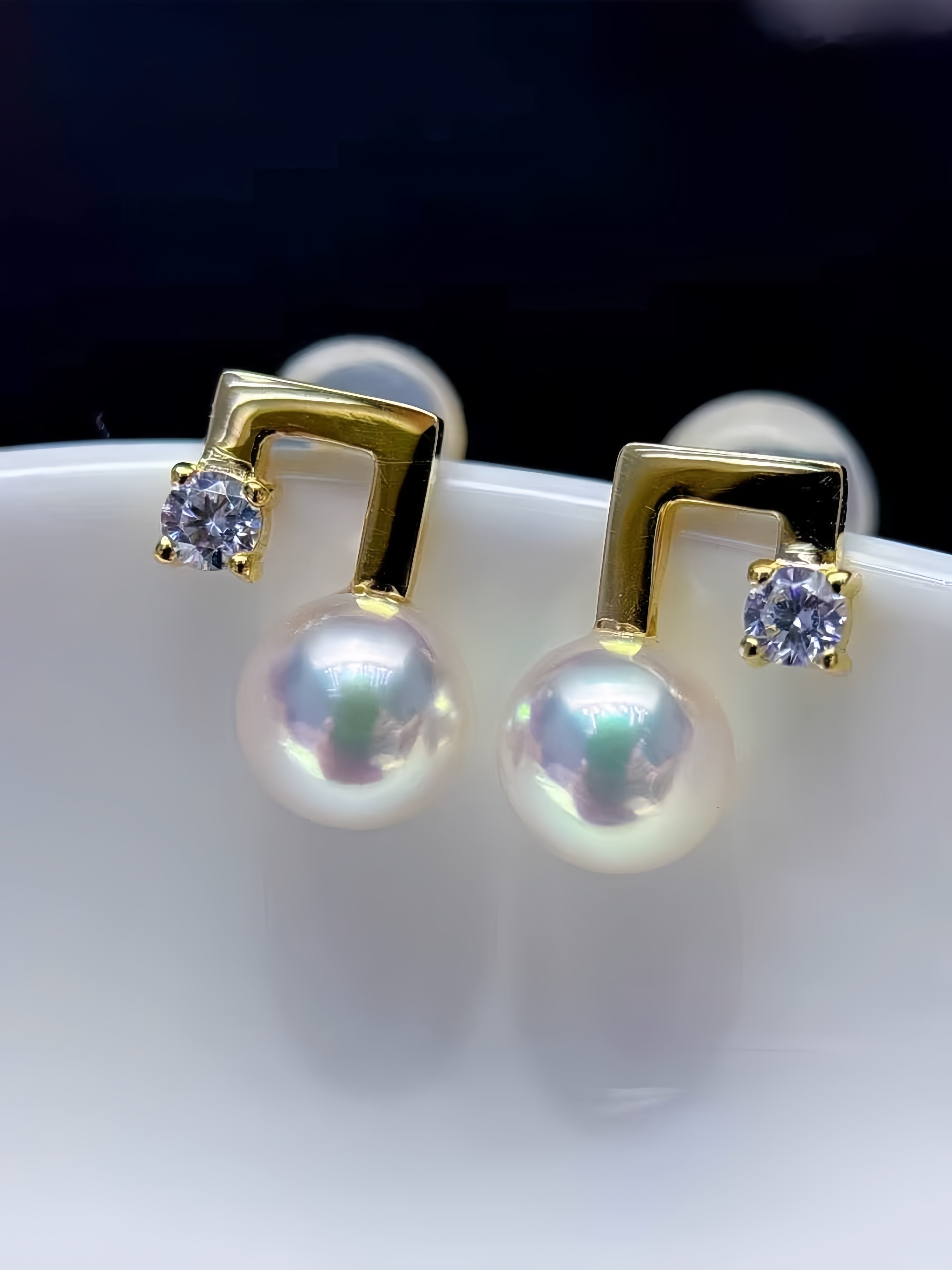 A pair of gold stud earrings featuring a small square-cut clear gemstone and a large round white pearl hanging below, displayed on a white surface against a dark background.