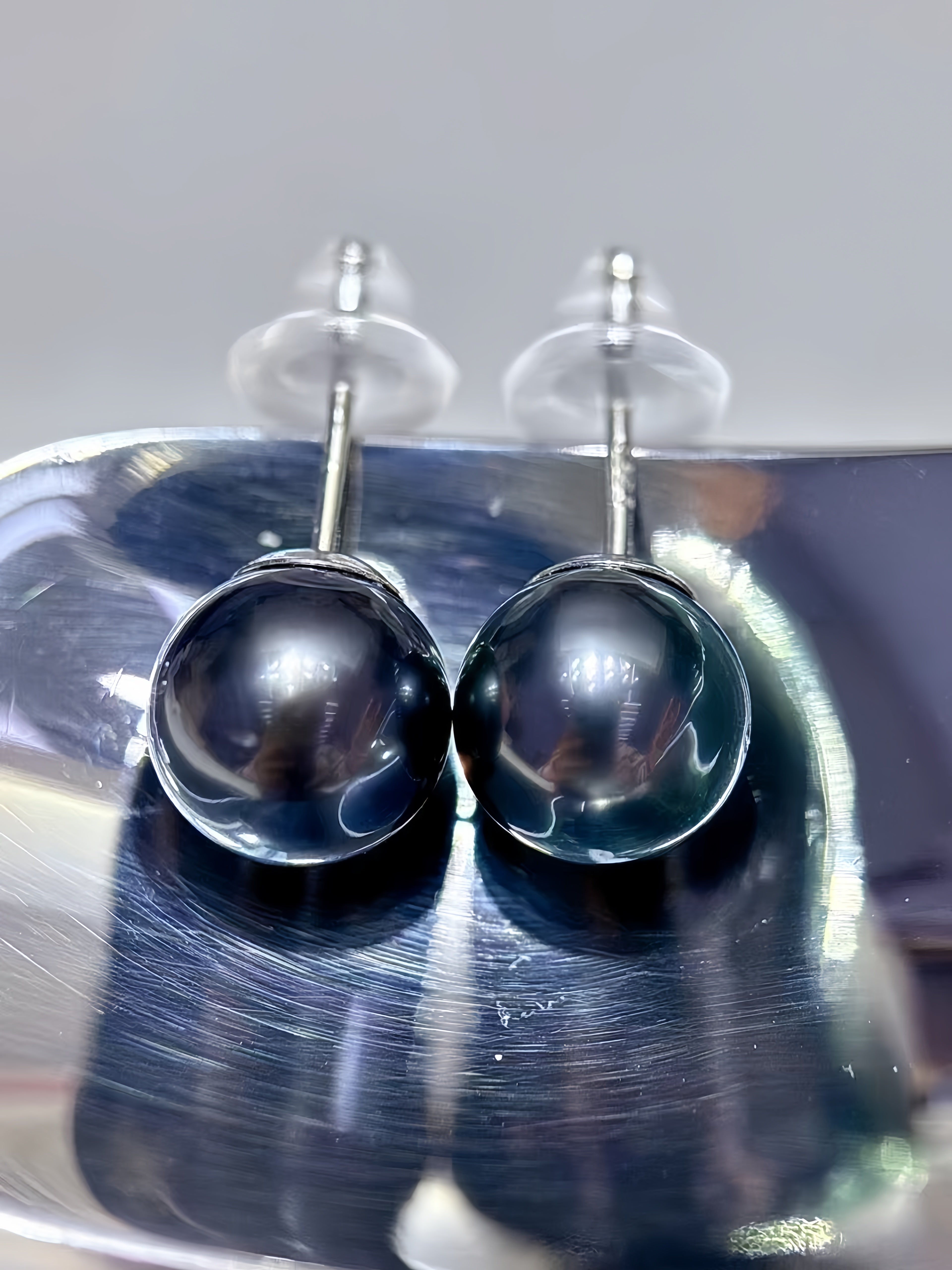 A pair of round black pearl stud earrings with silver posts and clear butterfly backings, resting on a reflective silver metal surface.