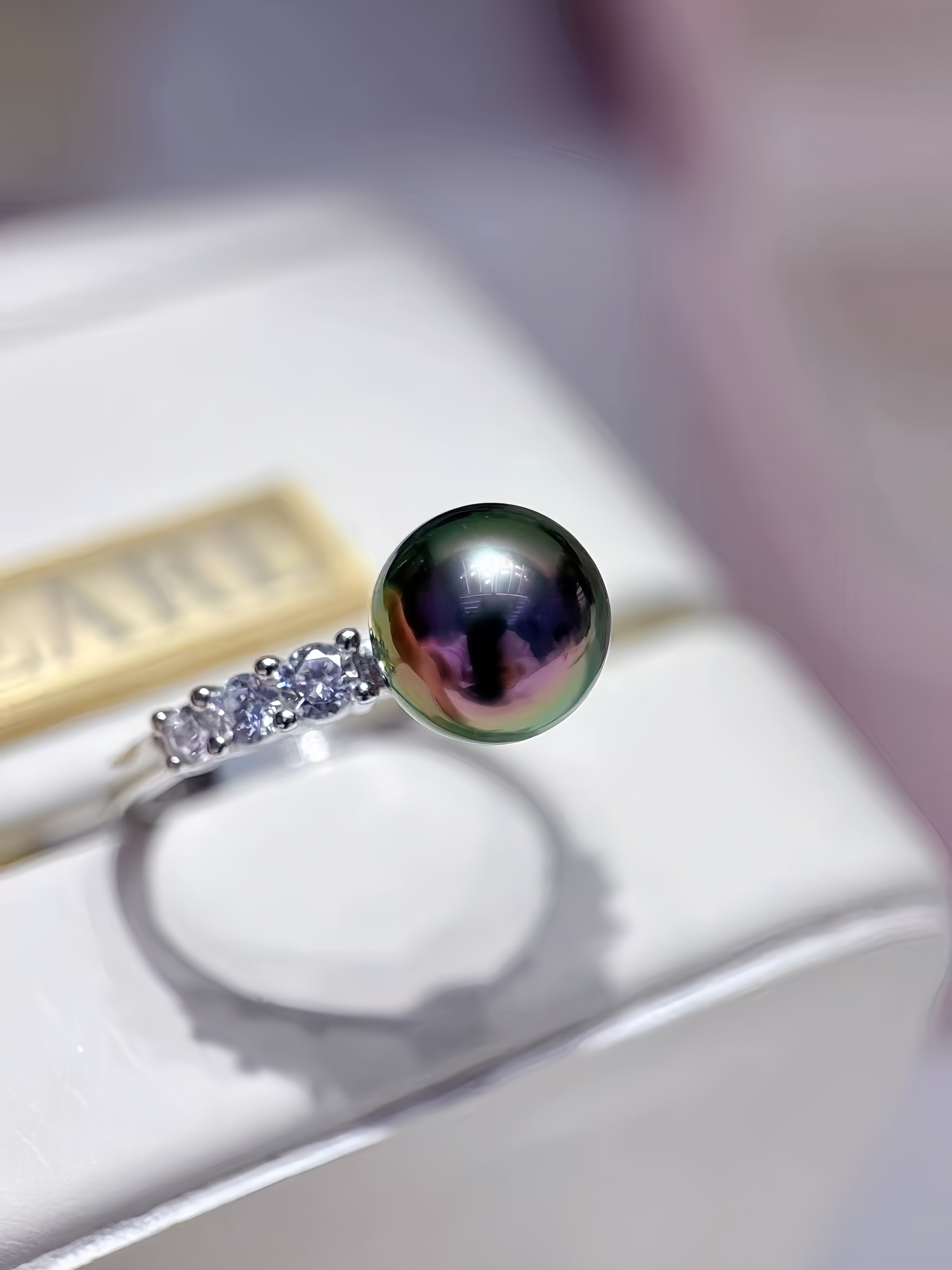 A silver ring featuring a large, dark green-black iridescent freshwater pearl and a row of small clear stones on the band, resting on a white jewelry display box.