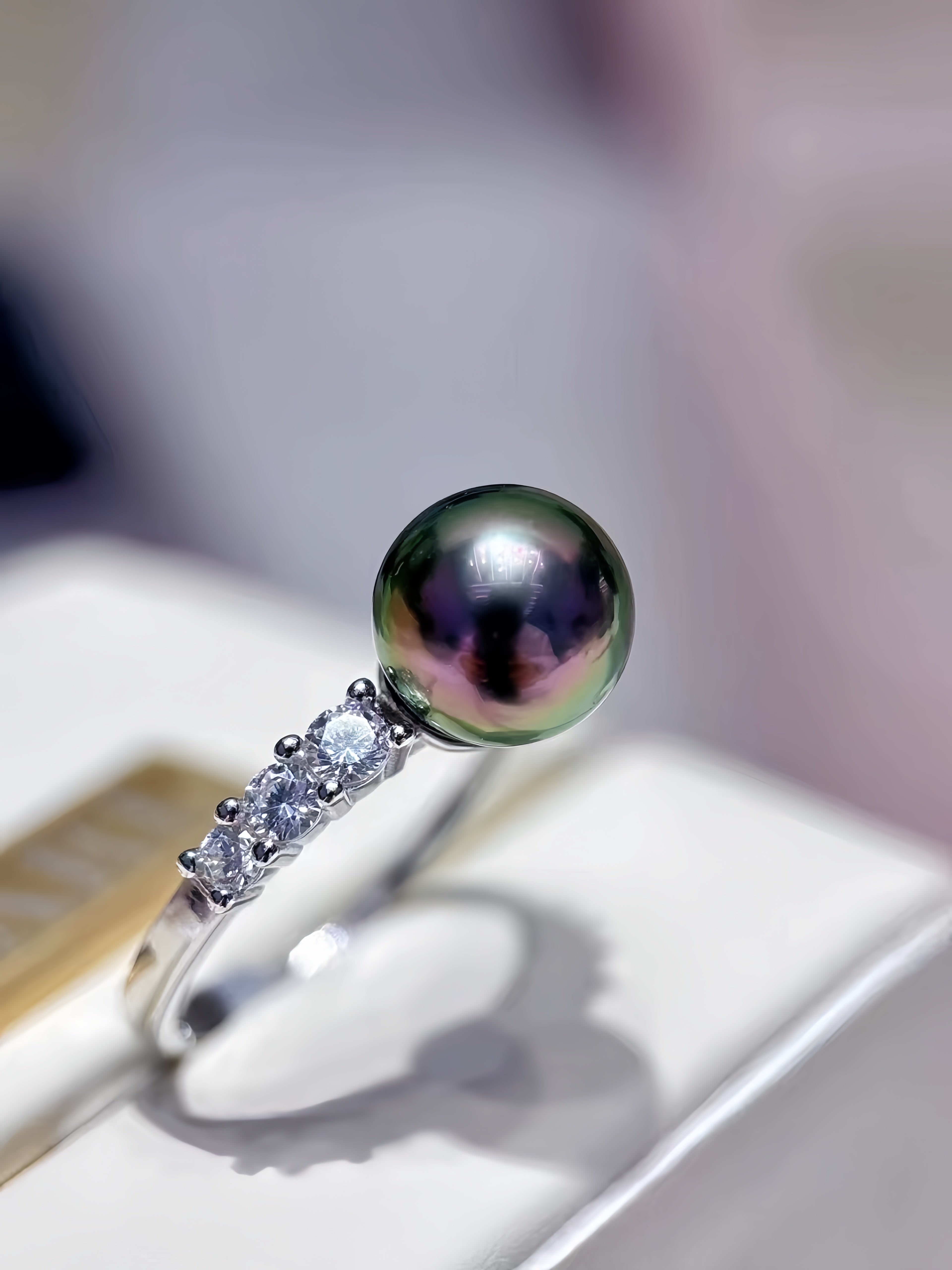 A silver ring featuring a large, round, iridescent green-black pearl on the top, with three small clear stones along the band, displayed on a white jewelry stand.