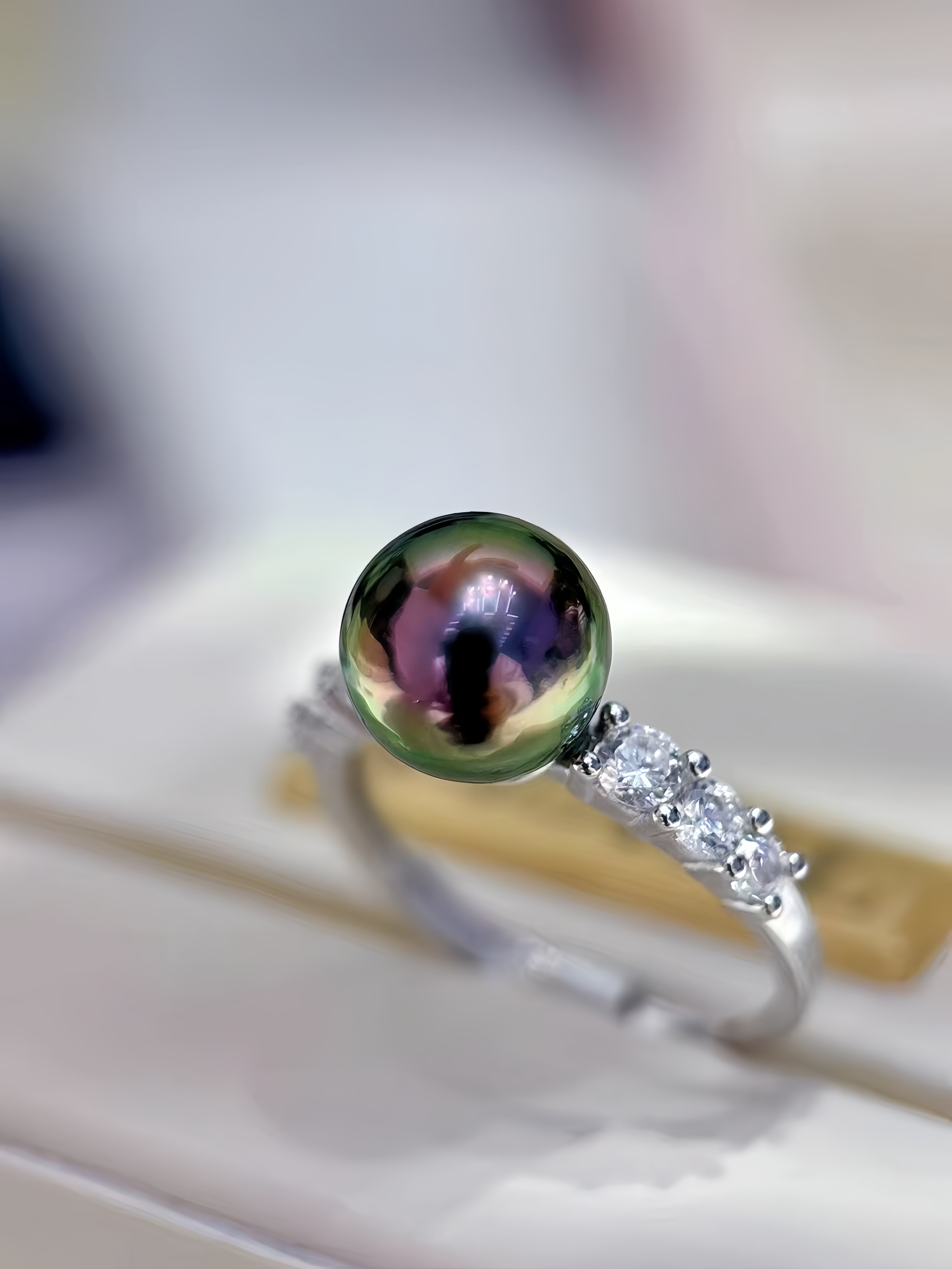 A silver ring featuring a large, round, iridescent green pearl with a reflective surface, accented by small clear stones on the band, resting on a white jewelry display box.