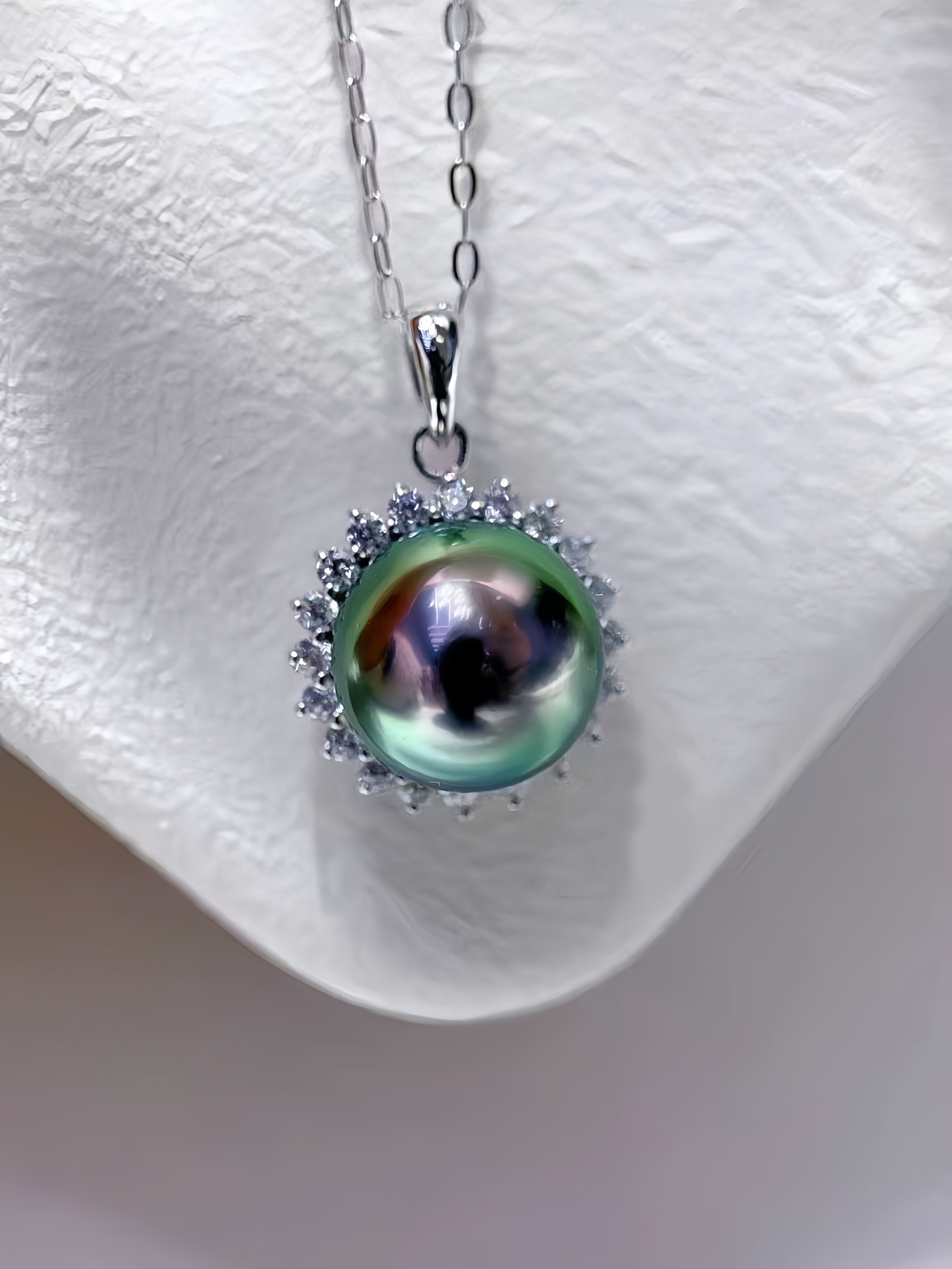 A silver pendant necklace featuring a large, iridescent green pearl surrounded by small clear stones, displayed on a textured white jewelry bust.