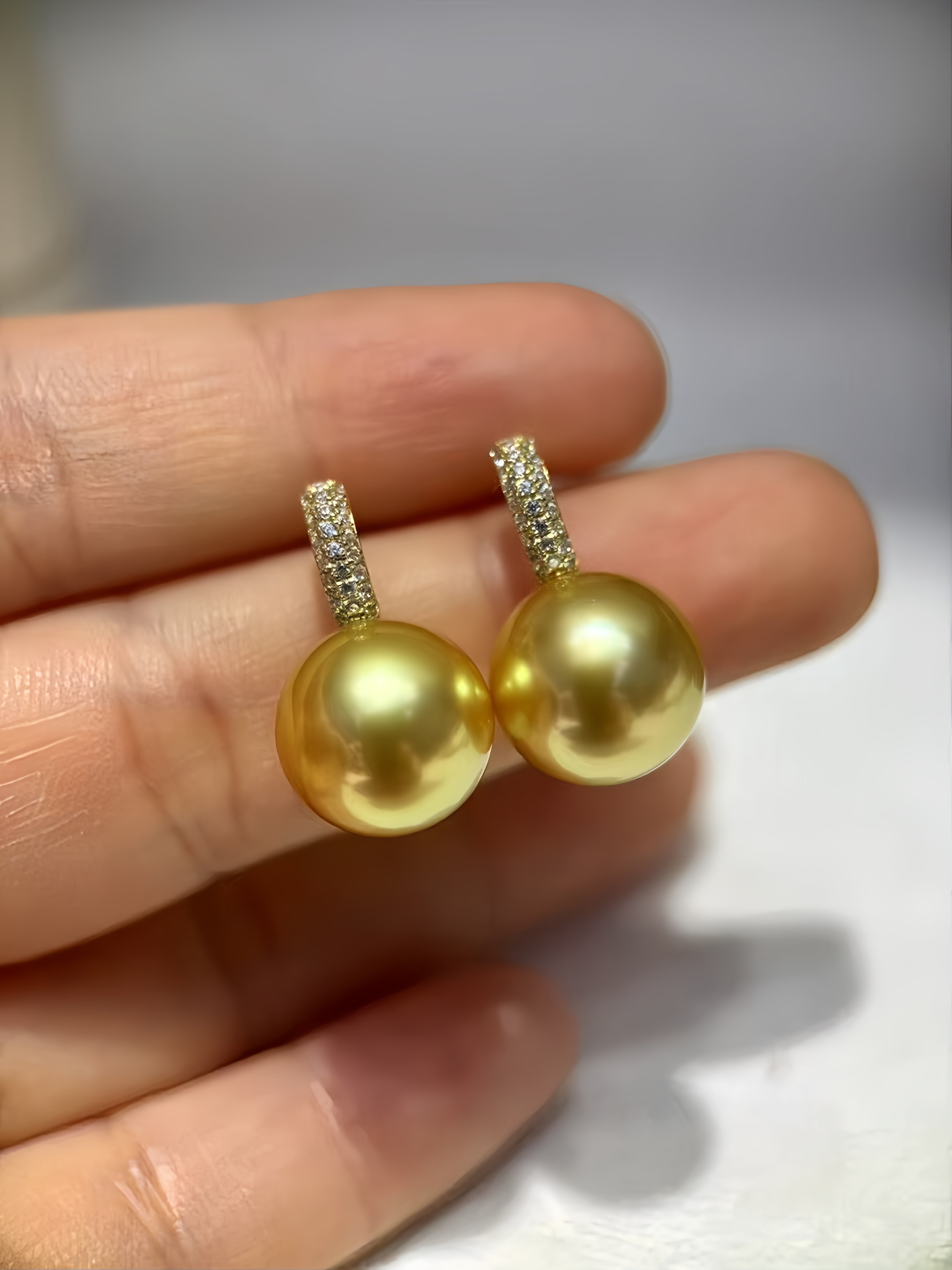 A pair of gold pearl drop earrings with small pavé diamond hoops, held between fingers to show scale.
