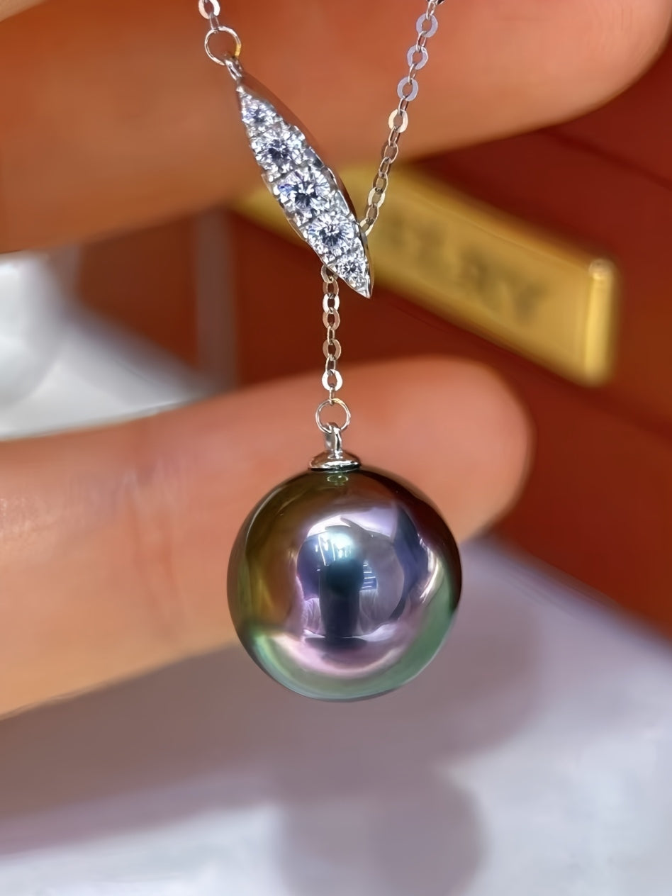 A close-up of a pendant necklace held between fingers, featuring a large iridescent dark pearl pendant hanging from a silver-toned chain with a pavé-set leaf-shaped charm. The background is softly blurred with warm brown tones.