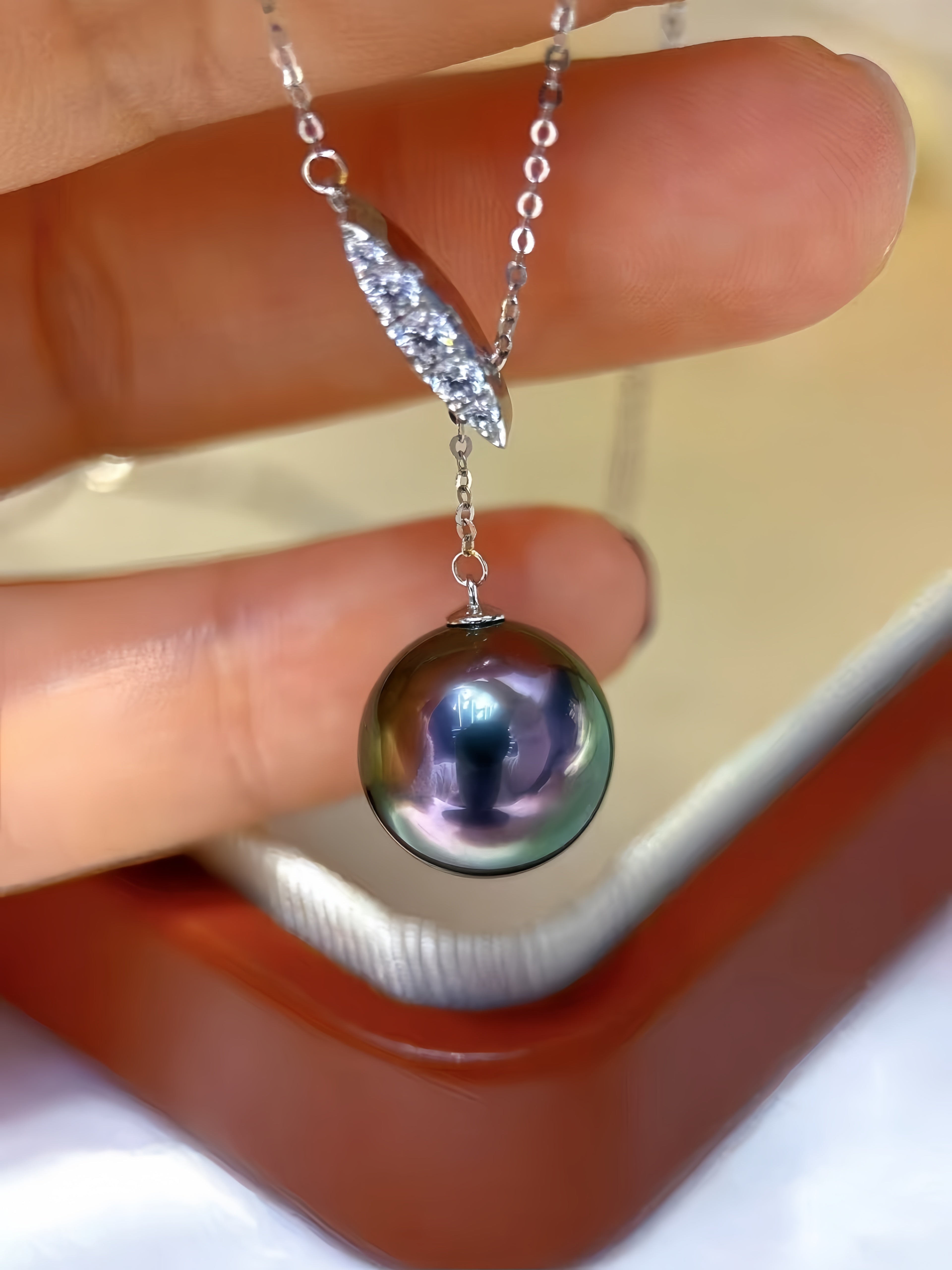 A close-up of a hand holding a silver pendant necklace with a large, iridescent dark pearl and a diamond-encrusted leaf-shaped connector.