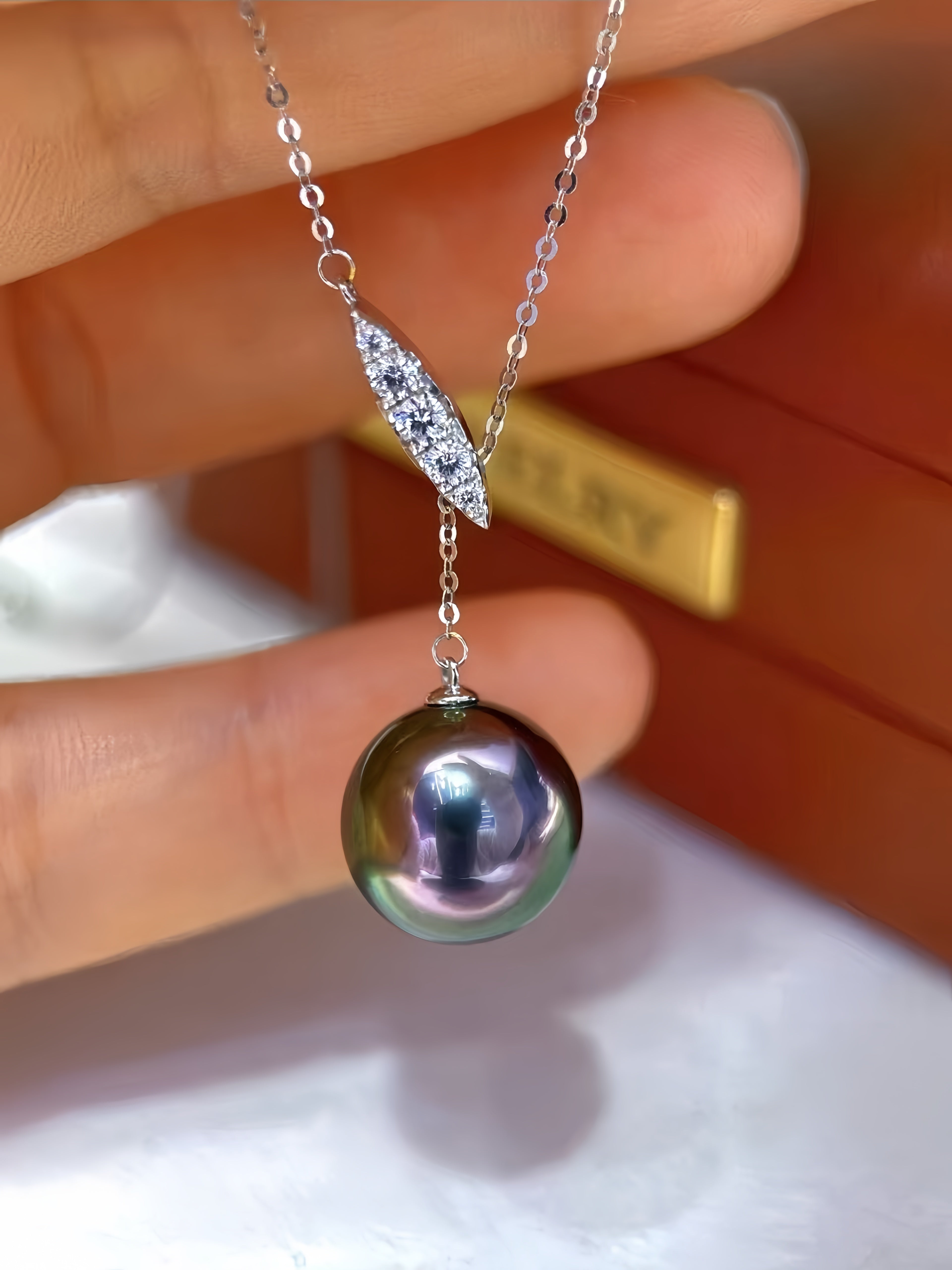 A close-up of a hand holding a silver pendant necklace featuring a large iridescent dark pearl and a diamond-accented leaf-shaped charm, with a blurred brown jewelry box in the background.