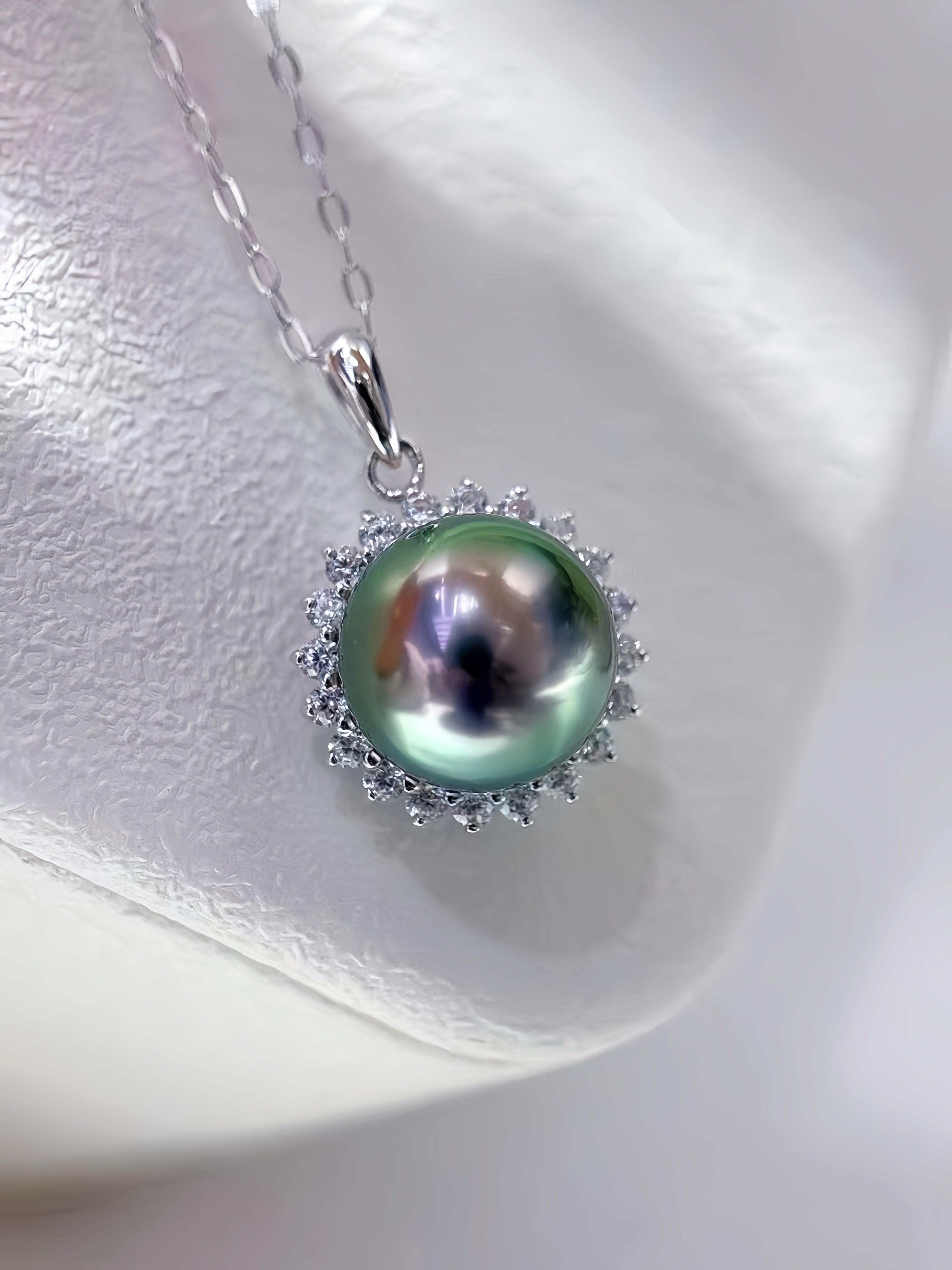 A silver pendant necklace featuring a large, round iridescent green pearl surrounded by a halo of small clear stones, displayed on a textured white surface.