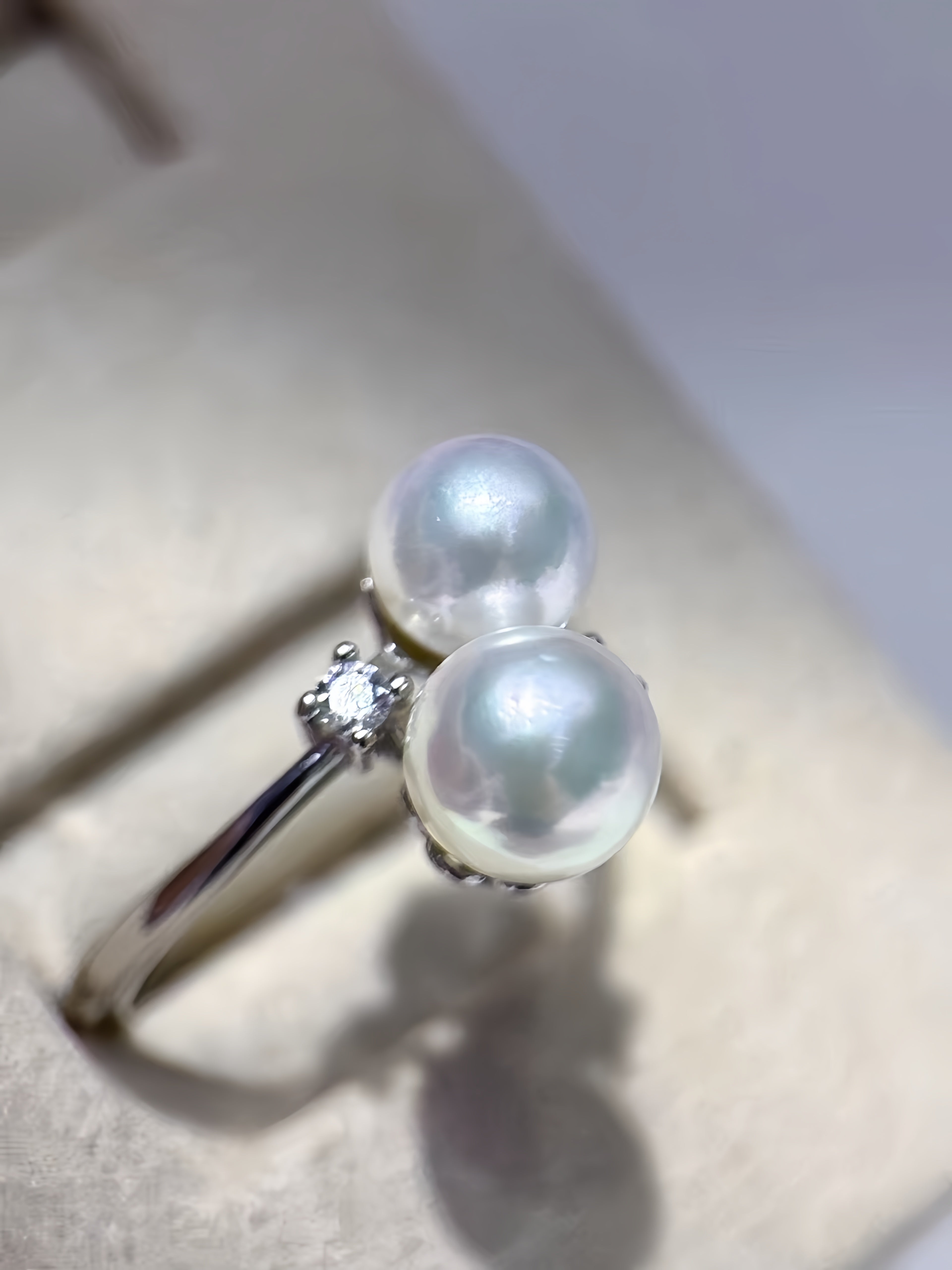 A silver ring featuring two round white pearls and a small clear accent stone, displayed on a beige jewelry stand.
