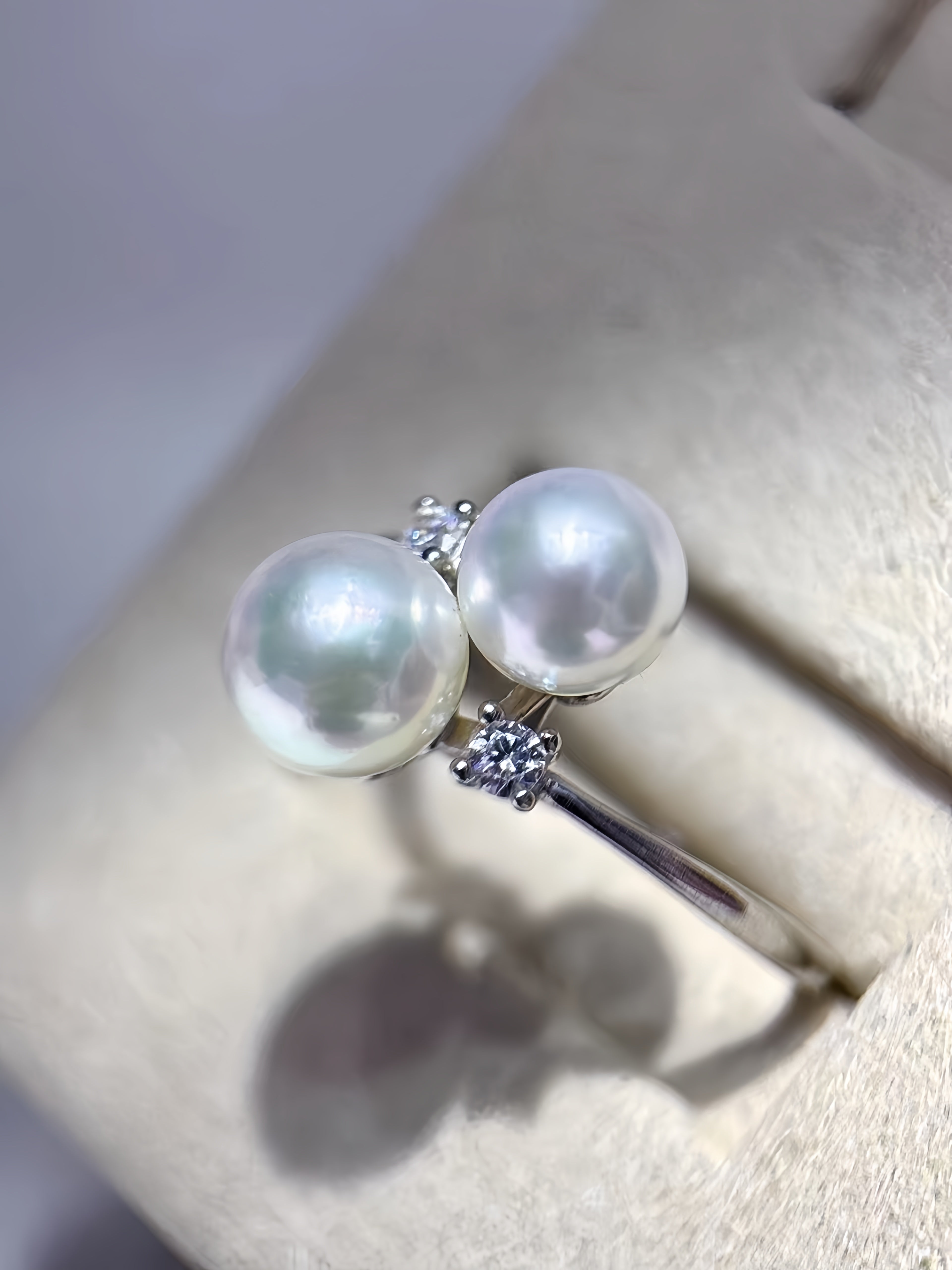 A silver ring featuring two large round white pearls and small clear accent stones, displayed on a beige jewelry stand.