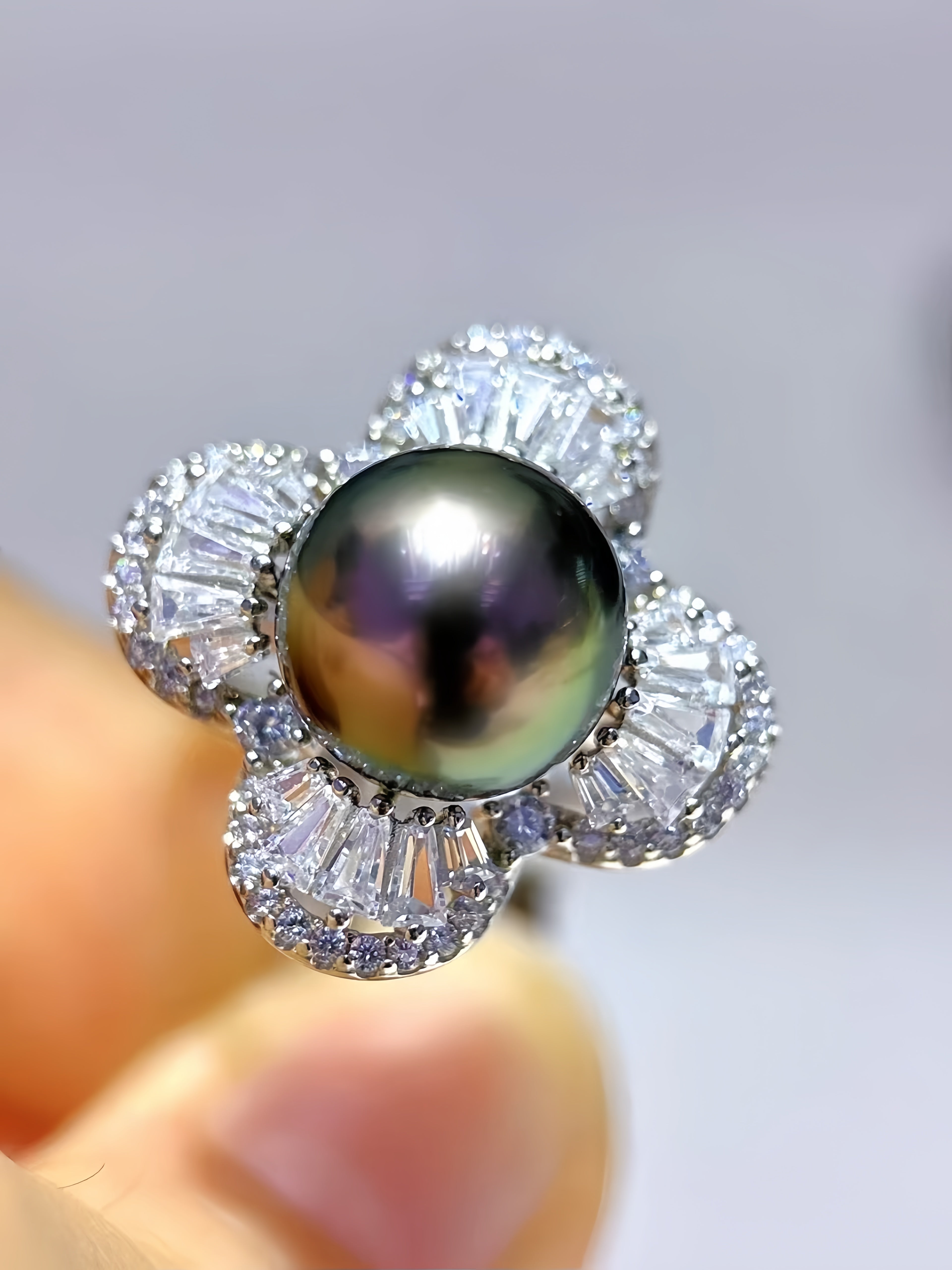 A close-up of a flower-shaped ring featuring a large iridescent dark pearl in the center, surrounded by four halo clusters of clear baguette-cut stones, held against a soft light gray background.
