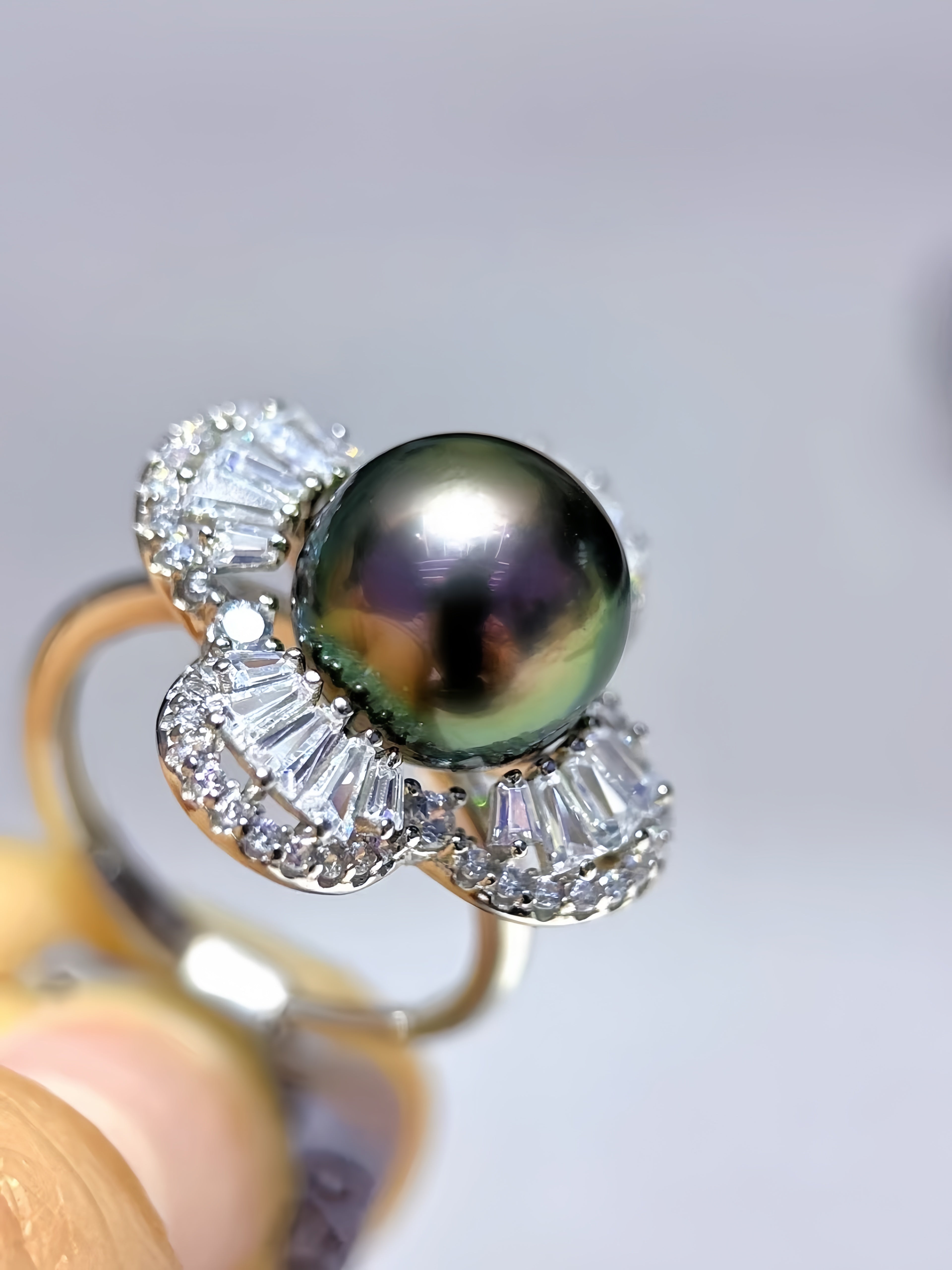A close-up of a ring held between fingers, featuring a large round iridescent dark green pearl in a silver-toned setting with clear baguette and round stones.