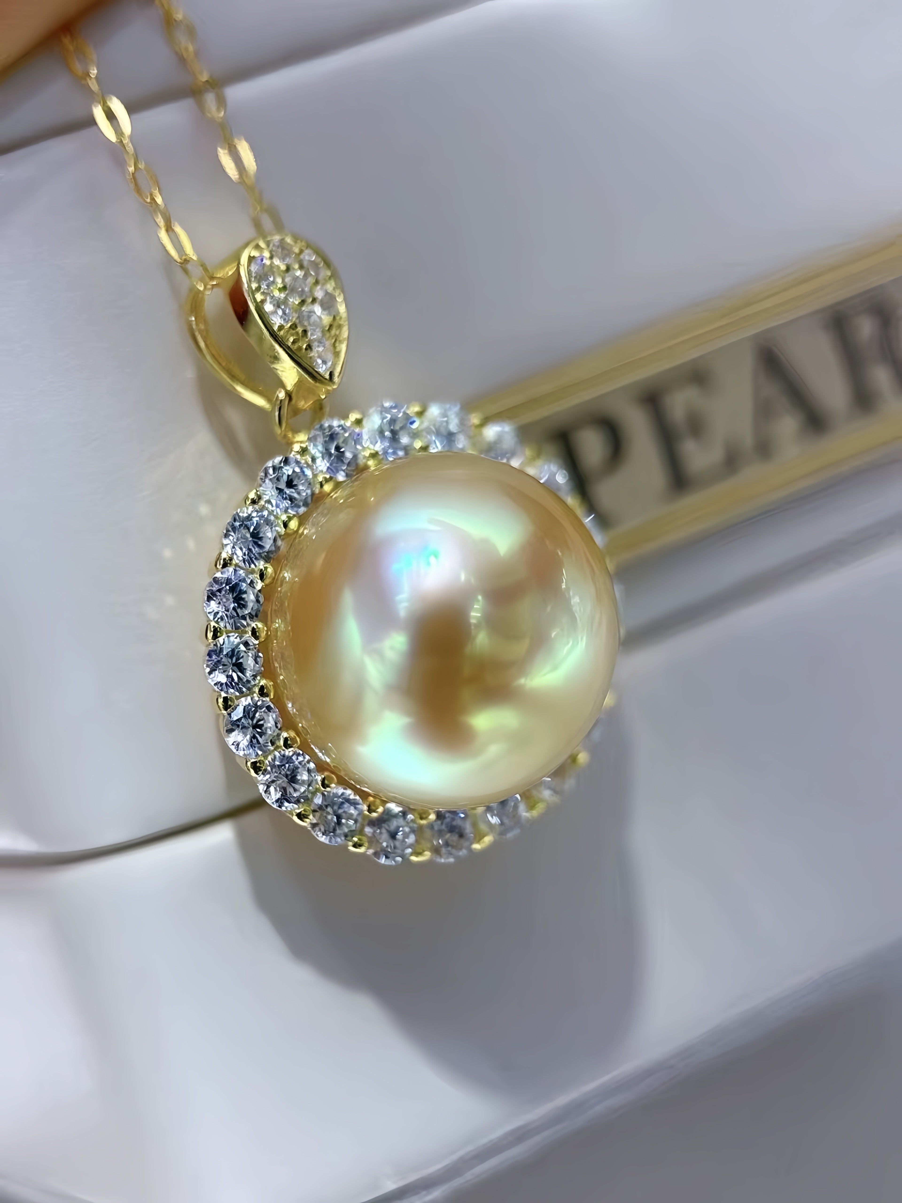 A gold pendant necklace featuring a large iridescent pearl surrounded by clear crystals, resting on a white jewelry box with a gold chain visible.