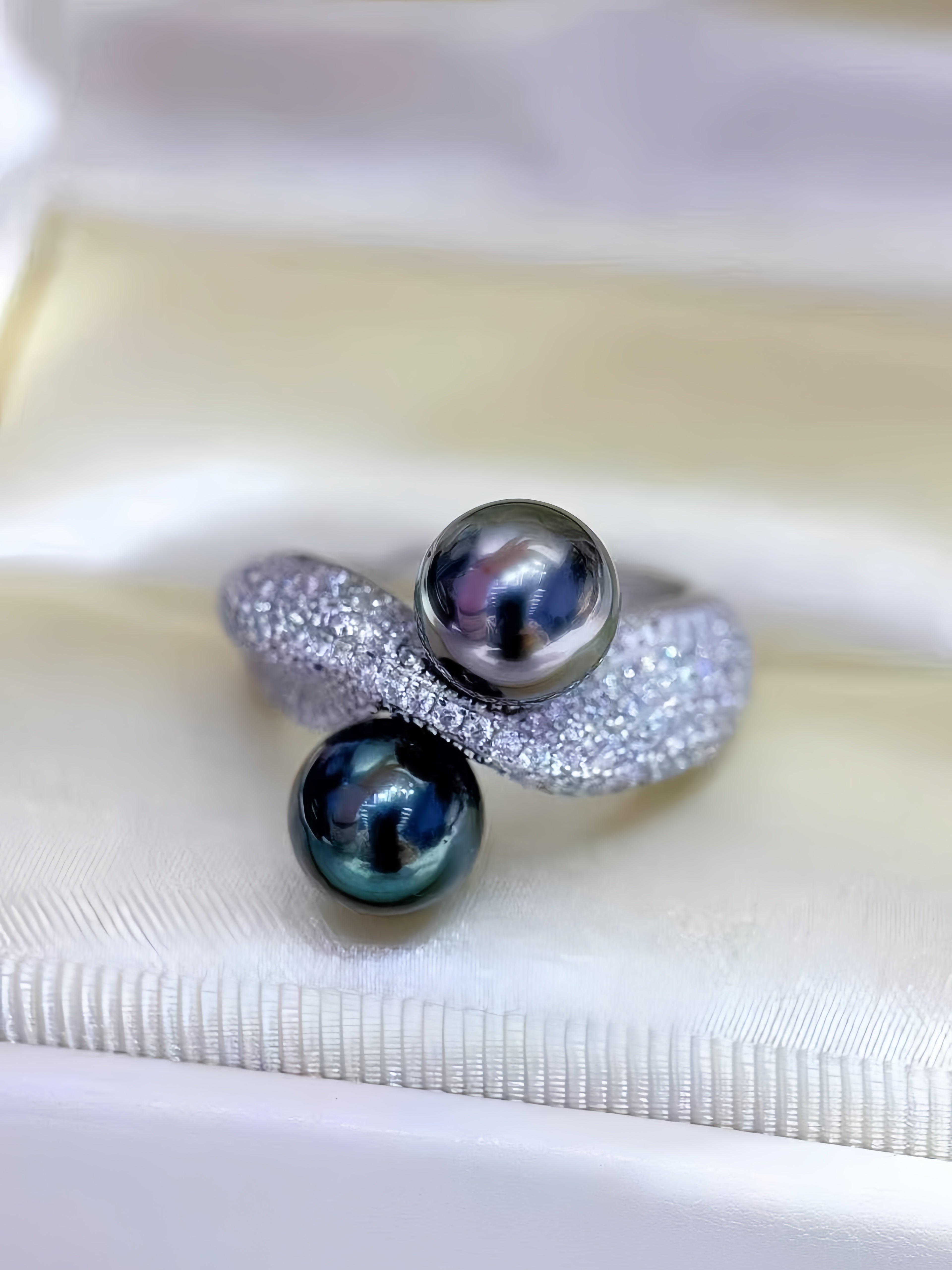 A silver ring featuring two dark pearls, one larger gray pearl and one smaller iridescent blue-green pearl, displayed on a white satin ring box.