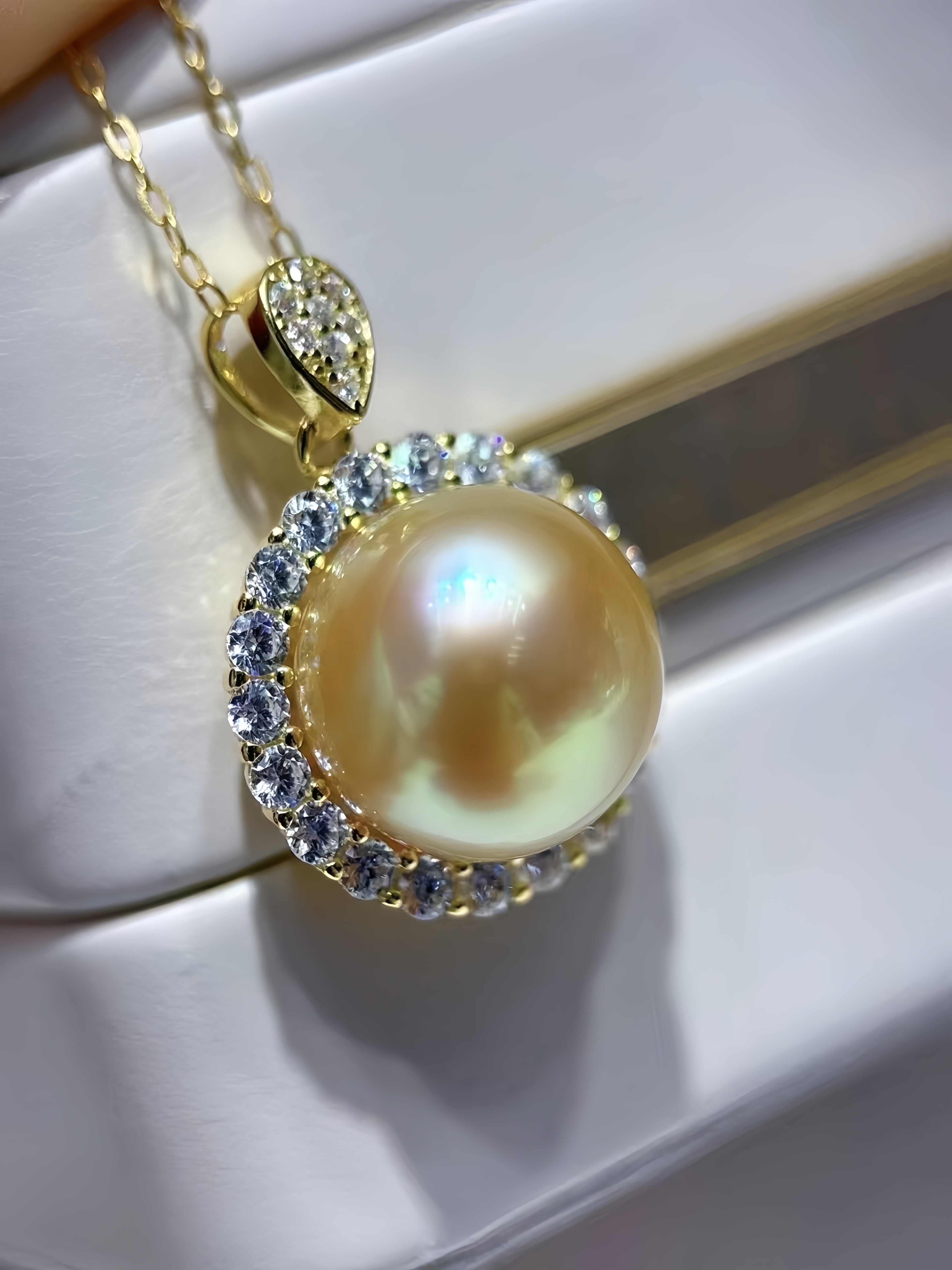 A gold pendant necklace featuring a large iridescent pearl surrounded by a halo of clear crystals, hanging from a gold chain on a white display surface.