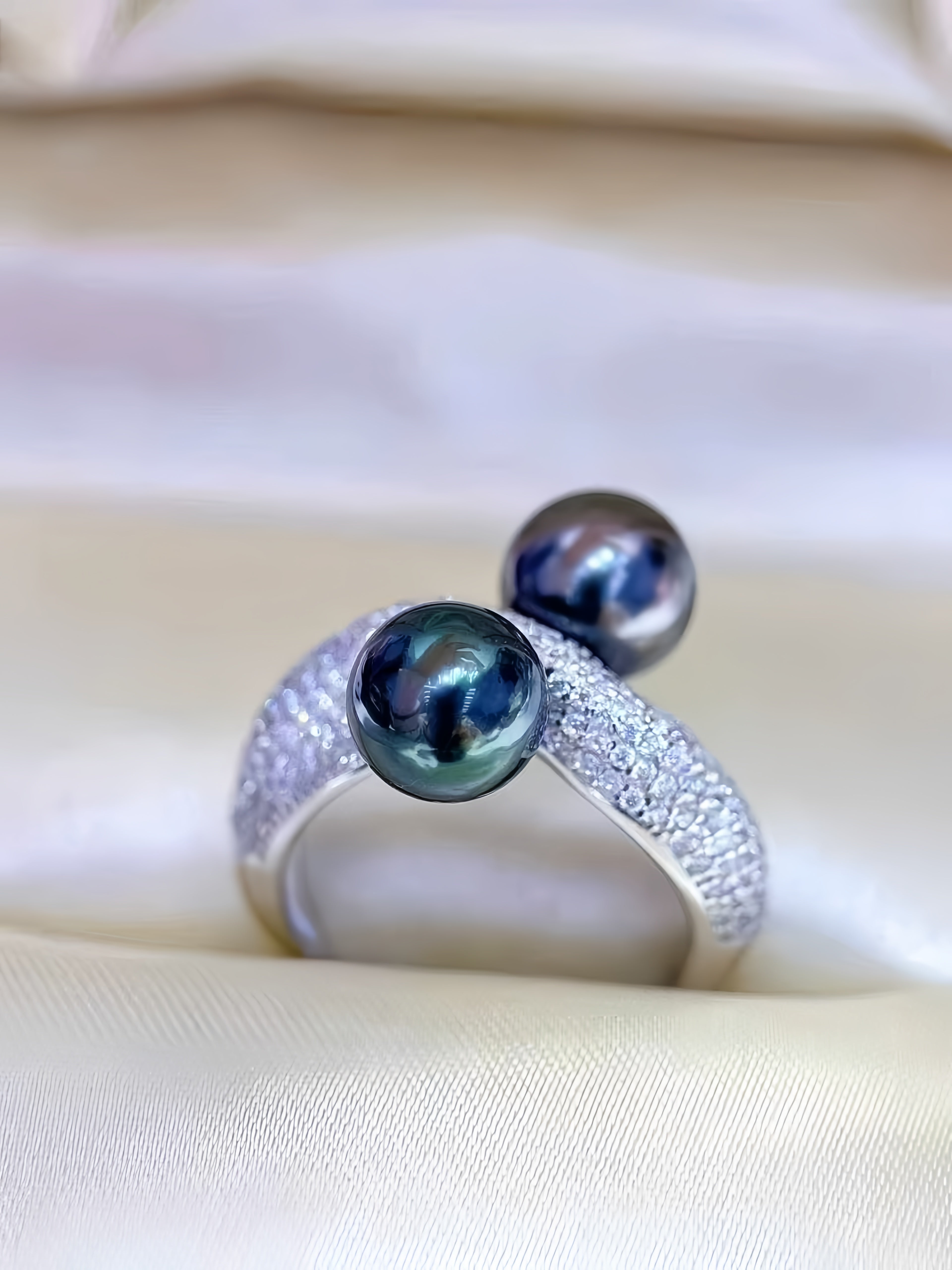 A silver ring featuring two dark, iridescent pearls and a pavé-set band, resting on a soft beige satin fabric background.